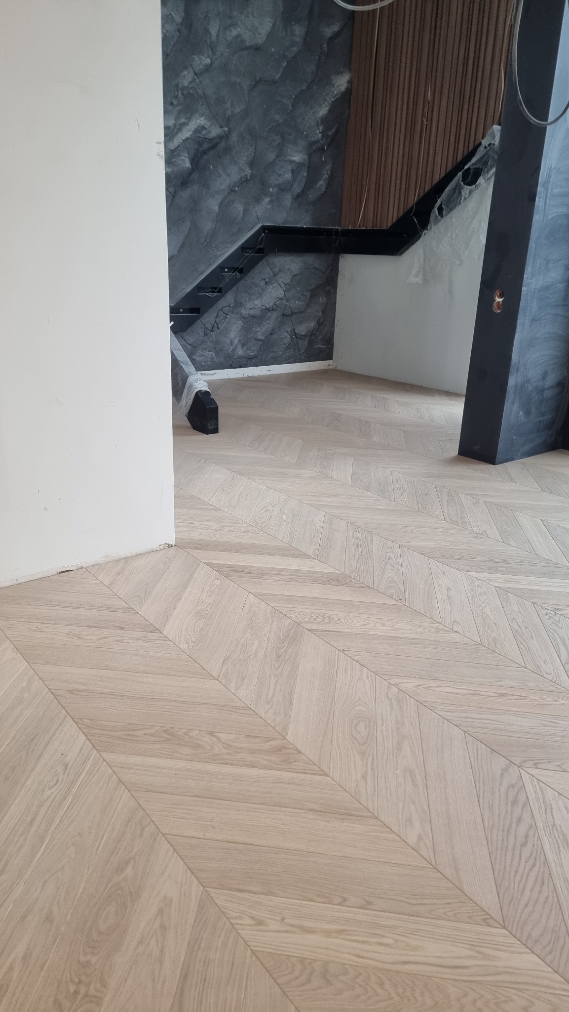 Jasny parkiet ułożony we wzór jodełki francuskiej w nowocześnie wykończonym wnętrzu. Widoczne fragmenty schodów i ściany z imitacją kamienia.