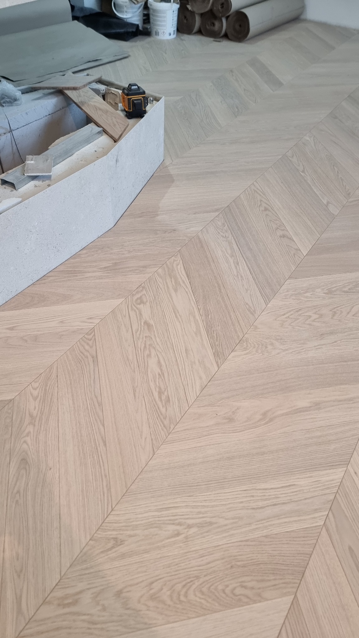 Ułożony parkiet w jodełkę francuską, jasne drewno. Na pierwszym planie listwa przypodłogowa i narzędzia. W tle rolki materiału wykończeniowego.