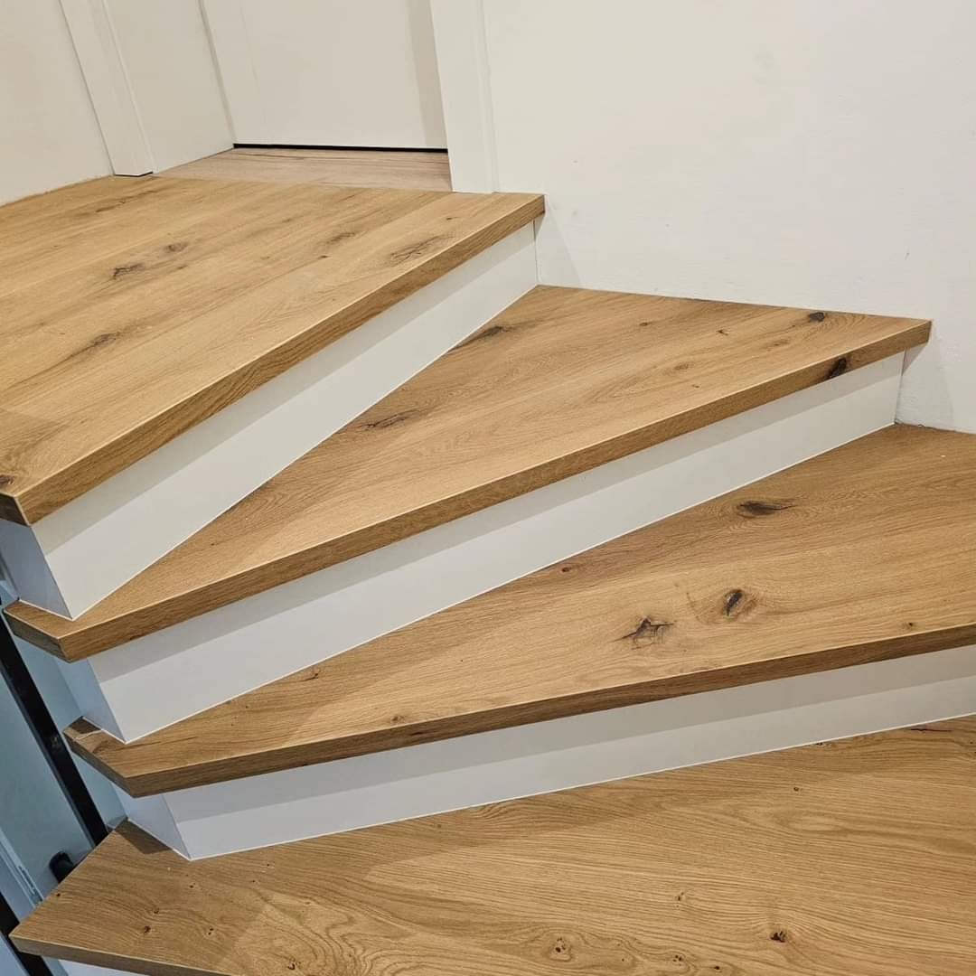 Drewniane schody o jasnym odcieniu, z białymi podstopnicami, ukazane z bliska. Widoczne słoje drewna i detale wykończenia. Minimalistyczny design.