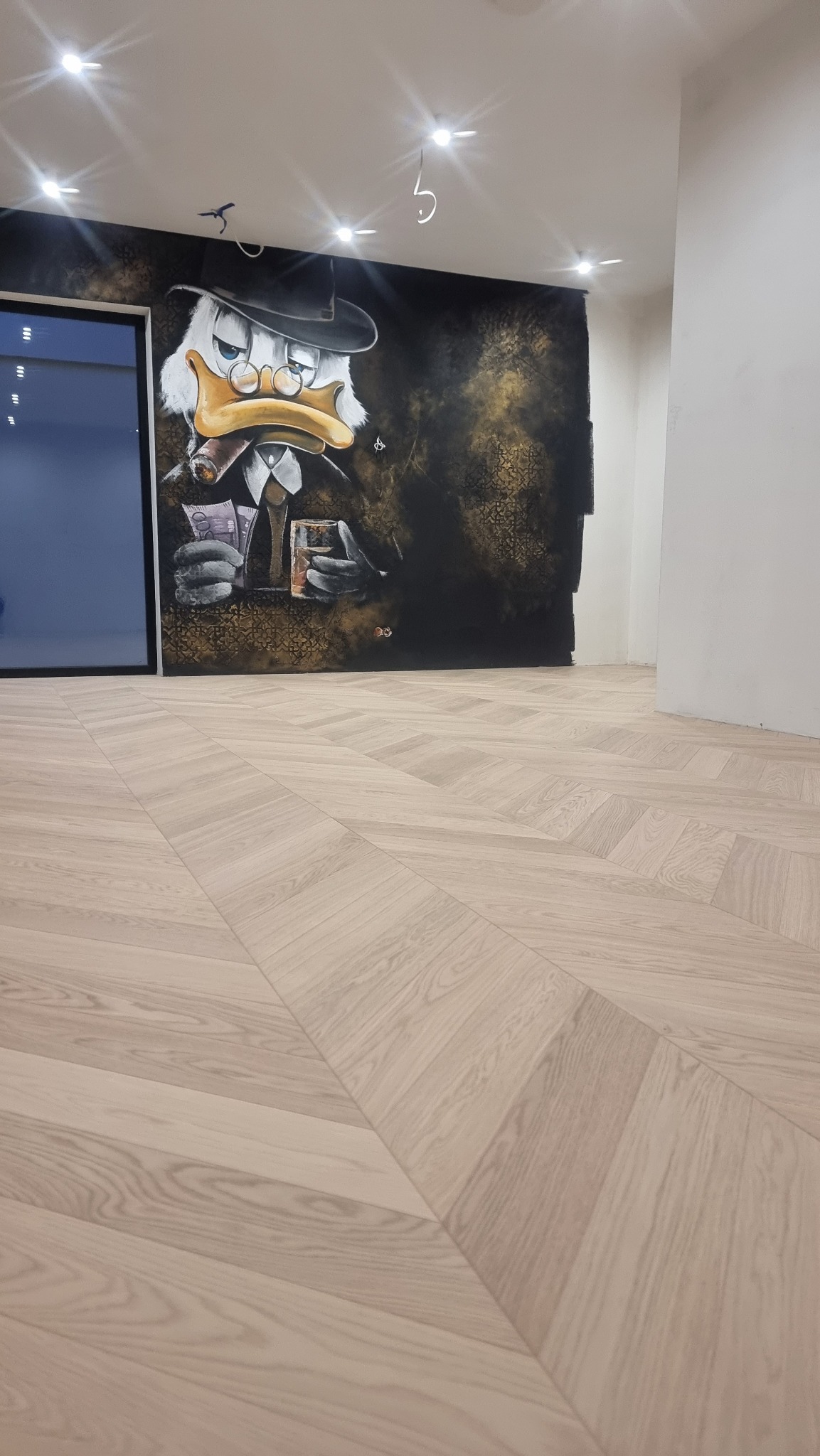 Jasne, drewniane panele ułożone we wzór jodełki francuskiej, z widocznym fragmentem ściany z muralem Kaczora Scrooge w tle. Nowoczesne wykończenie wnętrza.