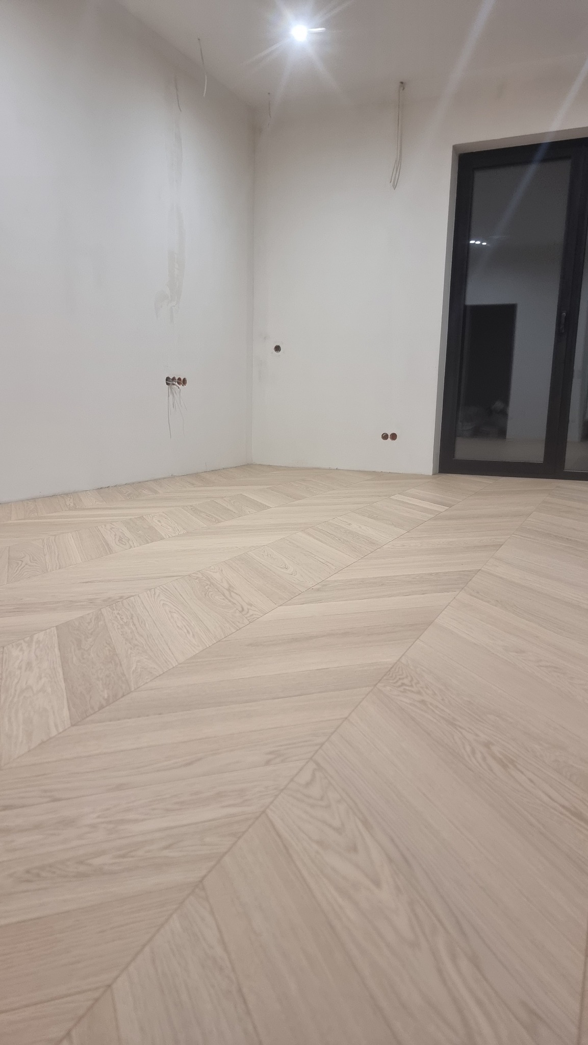 Jasna podłoga ułożona we wzór jodełki francuskiej w pokoju z białymi ścianami i czarną ramą drzwiową. Widoczne niedokończone gniazdka elektryczne.