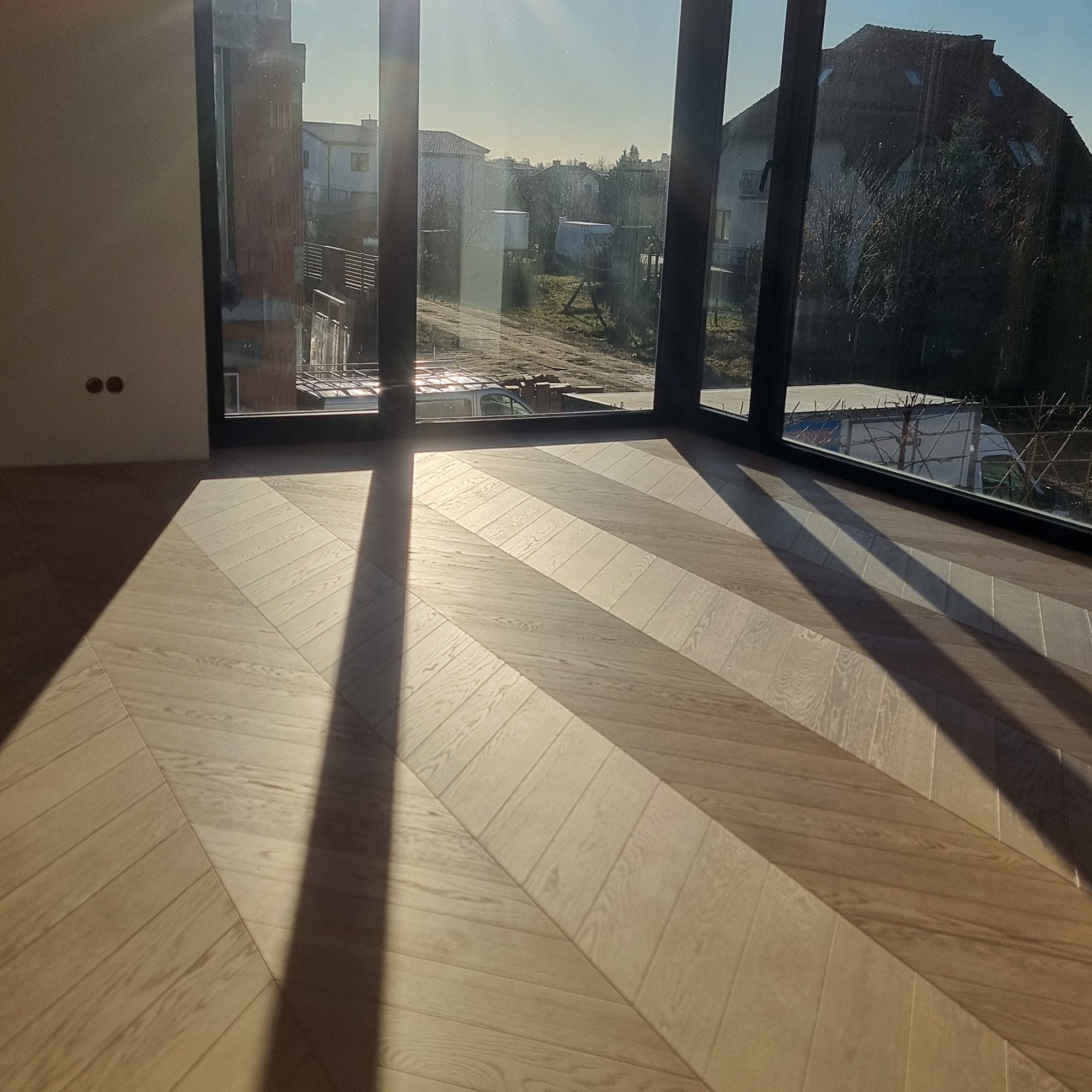 Jasna, drewniana podłoga ułożona w jodełkę, oświetlona promieniami słońca wpadającymi przez okno. Widok na budynki w tle.