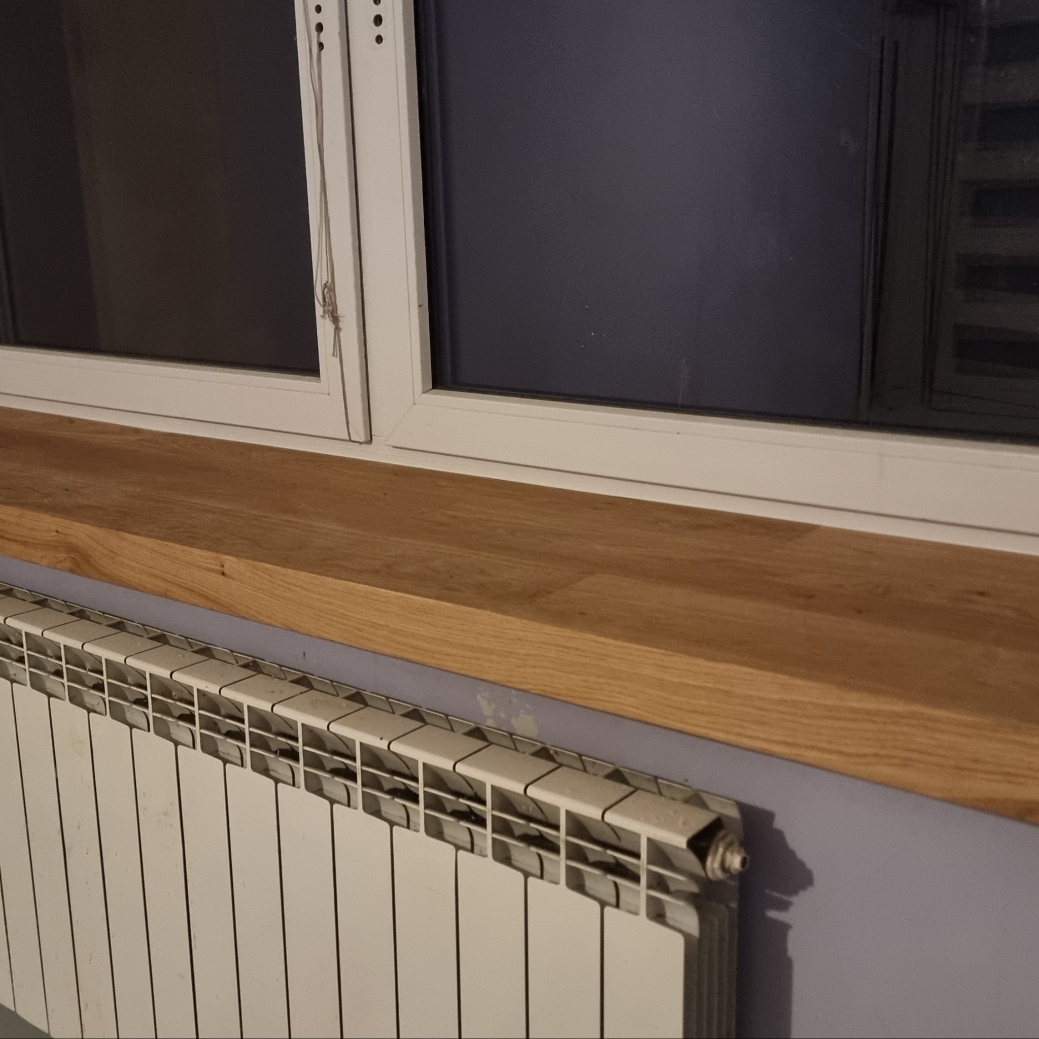 Zamontowany drewniany parapet pod oknem, z grzejnikiem poniżej, na tle fioletowej ściany. Widoczne detale wykończenia i naturalna faktura drewna.