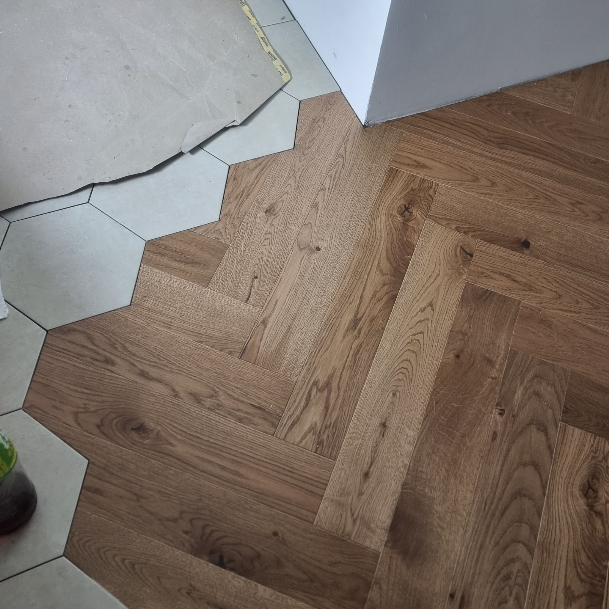 Parkiet ułożony we wzór jodełki, kontrastujący z szarymi, heksagonalnymi płytkami. Widoczne łączenie dwóch rodzajów podłóg. Oświetlenie naturalne.