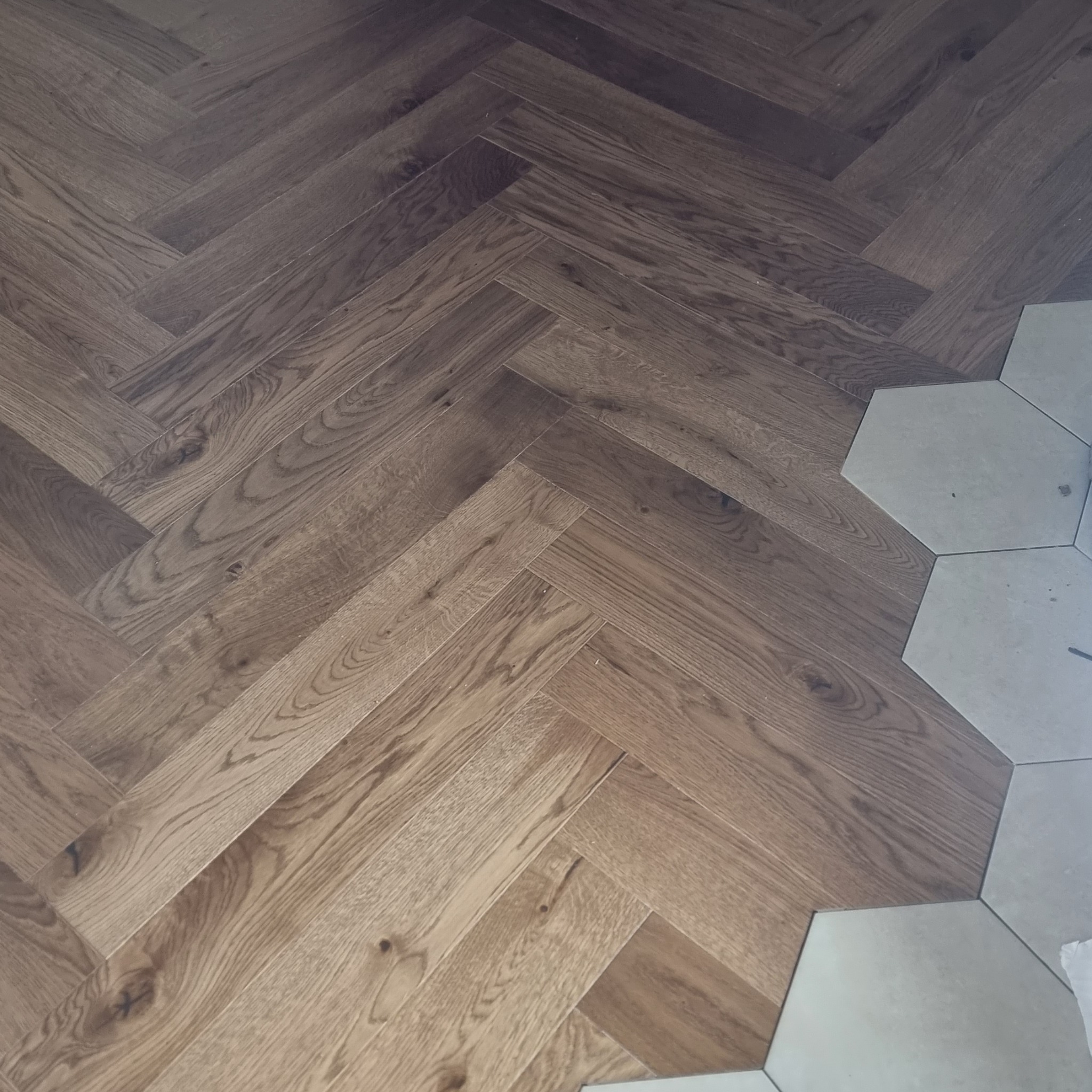 Parkiet ułożony we wzór jodełki francuskiej, kontrastujący z jasnymi, heksagonalnymi płytkami w rogu. Widoczne detale struktury drewna i fugi płytek.