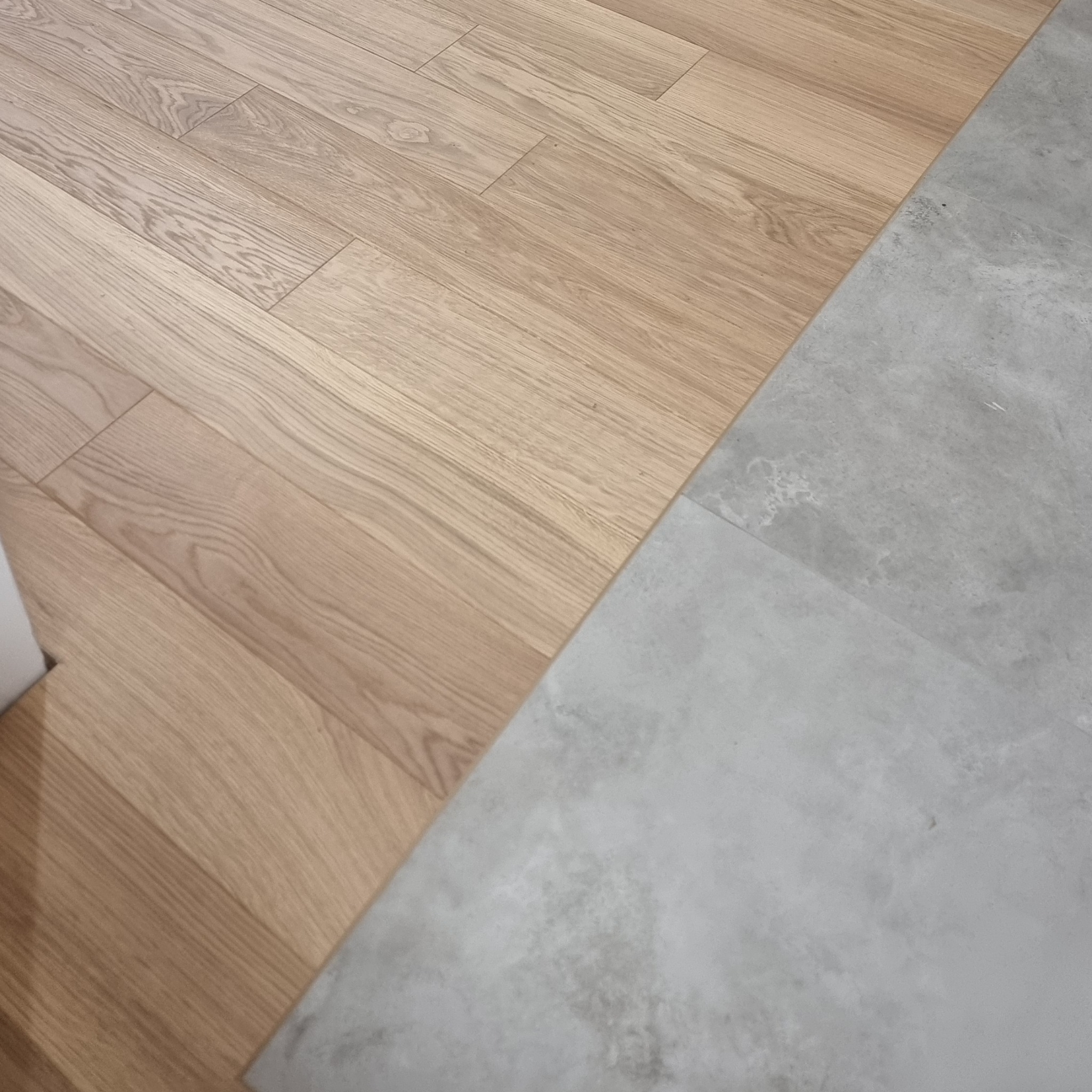 Połączenie podłogi z jasnego drewna i szarej, betonowej wylewki. Widoczne naturalne usłojenie drewna i faktura betonu, tworzące nowoczesny kontrast.