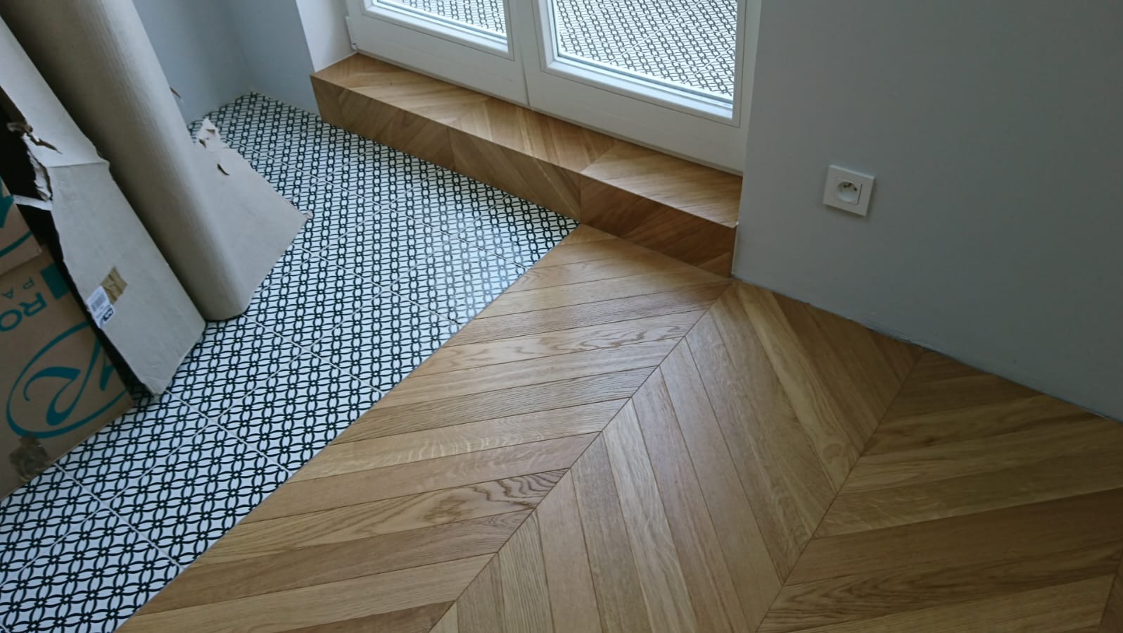 Parkiet ułożony we wzór jodełki francuskiej, kontrastujący z czarno-białą mozaiką przy oknie. Częściowo widoczne kartony z panelami. Gniazdko elektryczne na szarej ścianie.