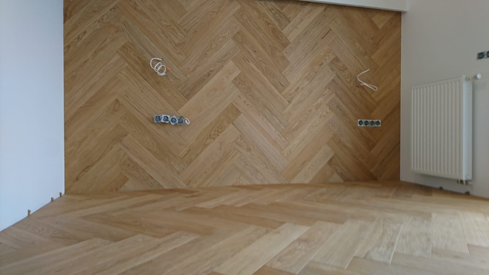Ułożony parkiet w jodełkę na podłodze i ścianie, widoczne gniazdka elektryczne i wystające kable, grzejnik. Jasne drewno, naturalne światło.
