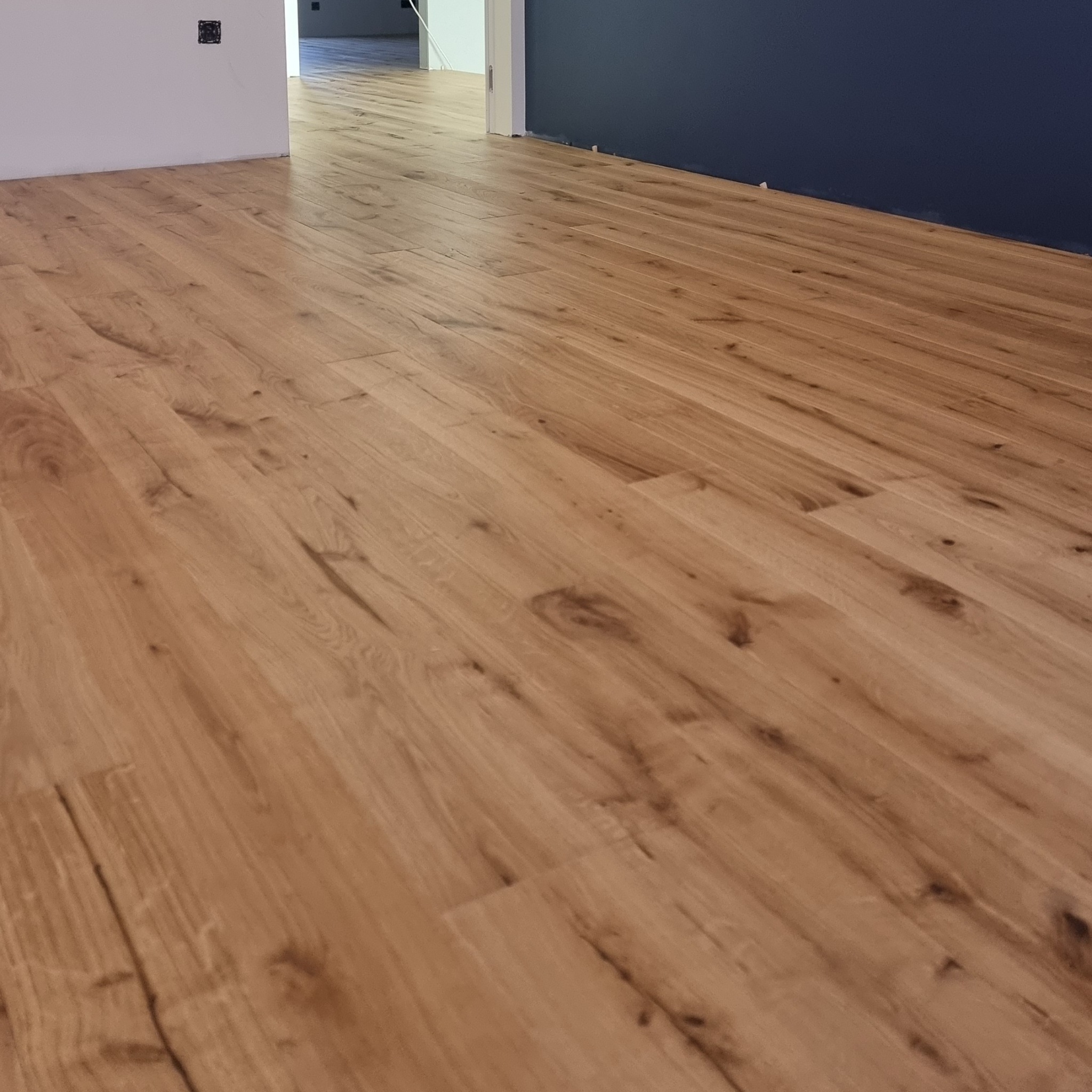 Ukończona podłoga z paneli w odcieniach dębu, ułożona w nowym mieszkaniu. Widoczne fragmenty ścian w kolorze białym i granatowym. Perspektywa z narożnika pokoju.