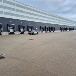 Libertad Robert Szydłowski - Długi budynek magazynowy z rampami załadunkowymi, biały pickup na placu. Pracownik w kamizelce przy rampie nr 54. Widok na szary plac z kostki brukowej.