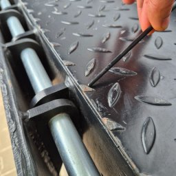 Libertad Robert Szydłowski - Czyszczenie metalowej konstrukcji z wzorem łezkowym. Palec wskazujący na rdzę, narzędzie w dłoni. Widoczne ślady korozji na czarnej powierzchni.