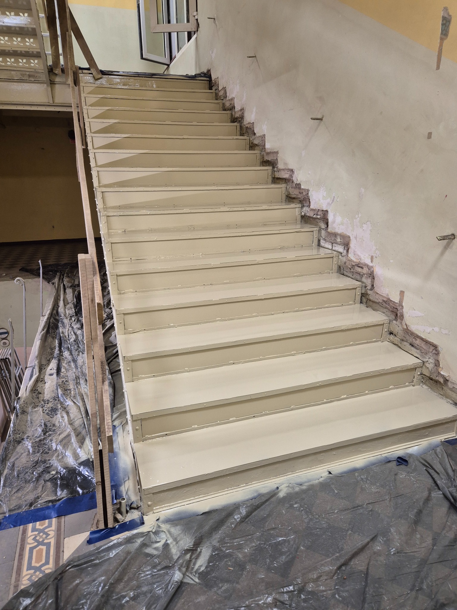 Malowane schody metalowe w trakcie renowacji. Widoczne zabezpieczenia podłoża folią malarską i niedomalowane fragmenty ściany. Ujęcie z dołu schodów.