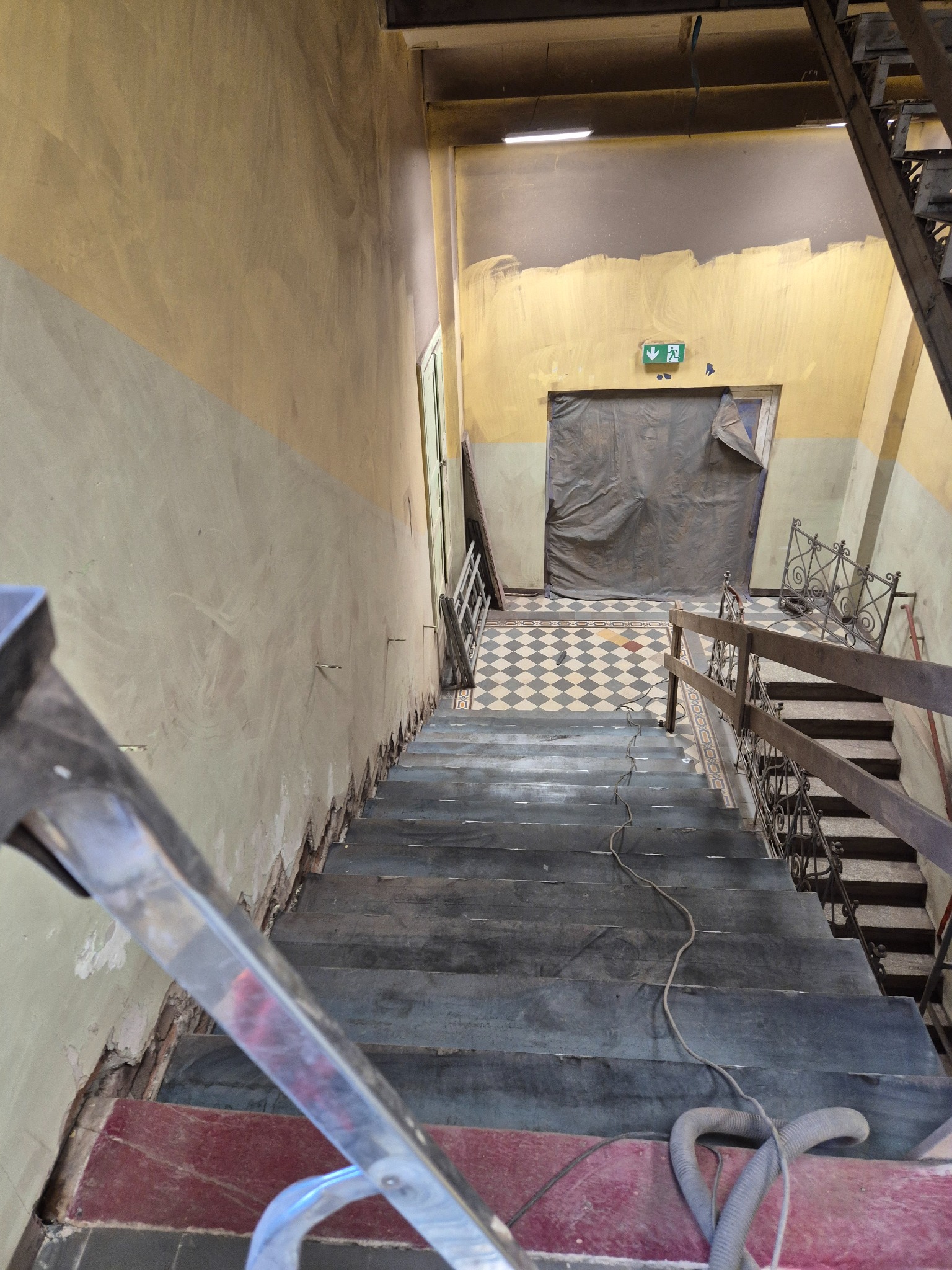 Klatka schodowa w trakcie remontu: widoczne uszkodzenia ścian, stare schody, zabezpieczone drzwi. Prace renowacyjne w budynku.