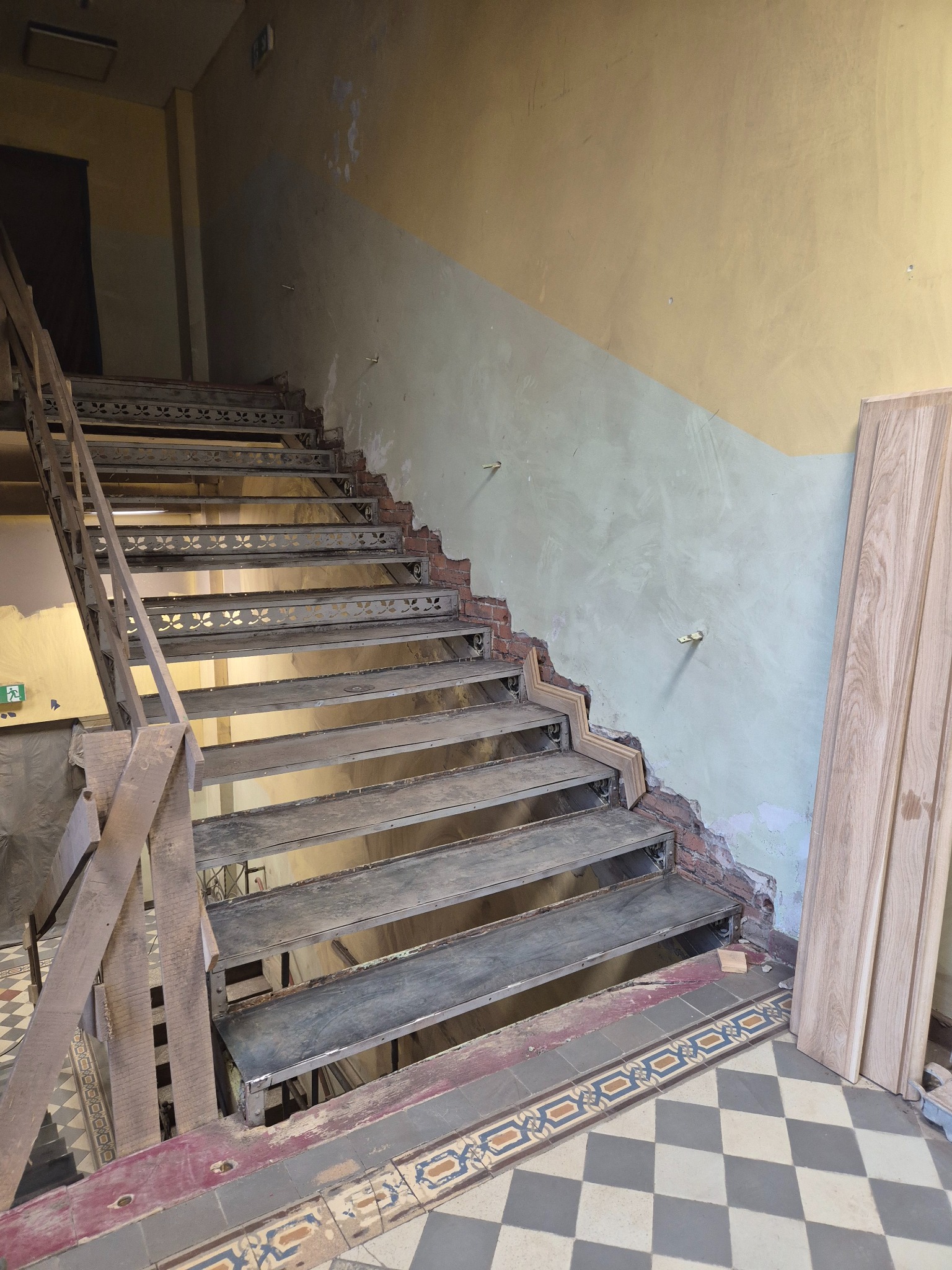 Metalowe schody w trakcie renowacji, widoczne elementy konstrukcyjne i przygotowane drewniane panele. Uwagę zwracają dekoracyjne detale na stopniach i wzorzysta podłoga.