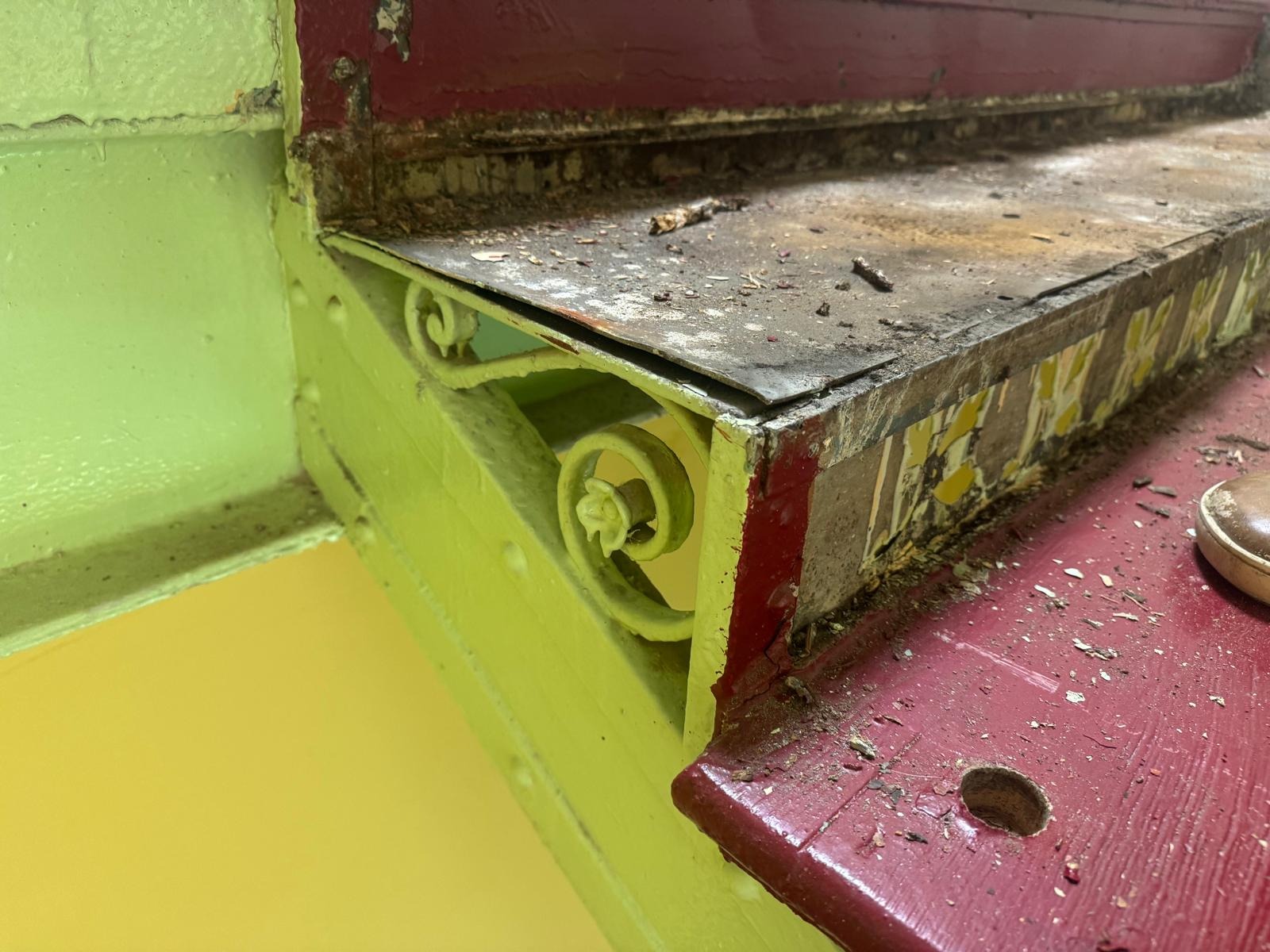 Zniszczone schody z metalową, dekoracyjną balustradą w trakcie renowacji. Widoczne ślady starej farby i zabrudzenia. Fragment buta w prawym dolnym rogu.