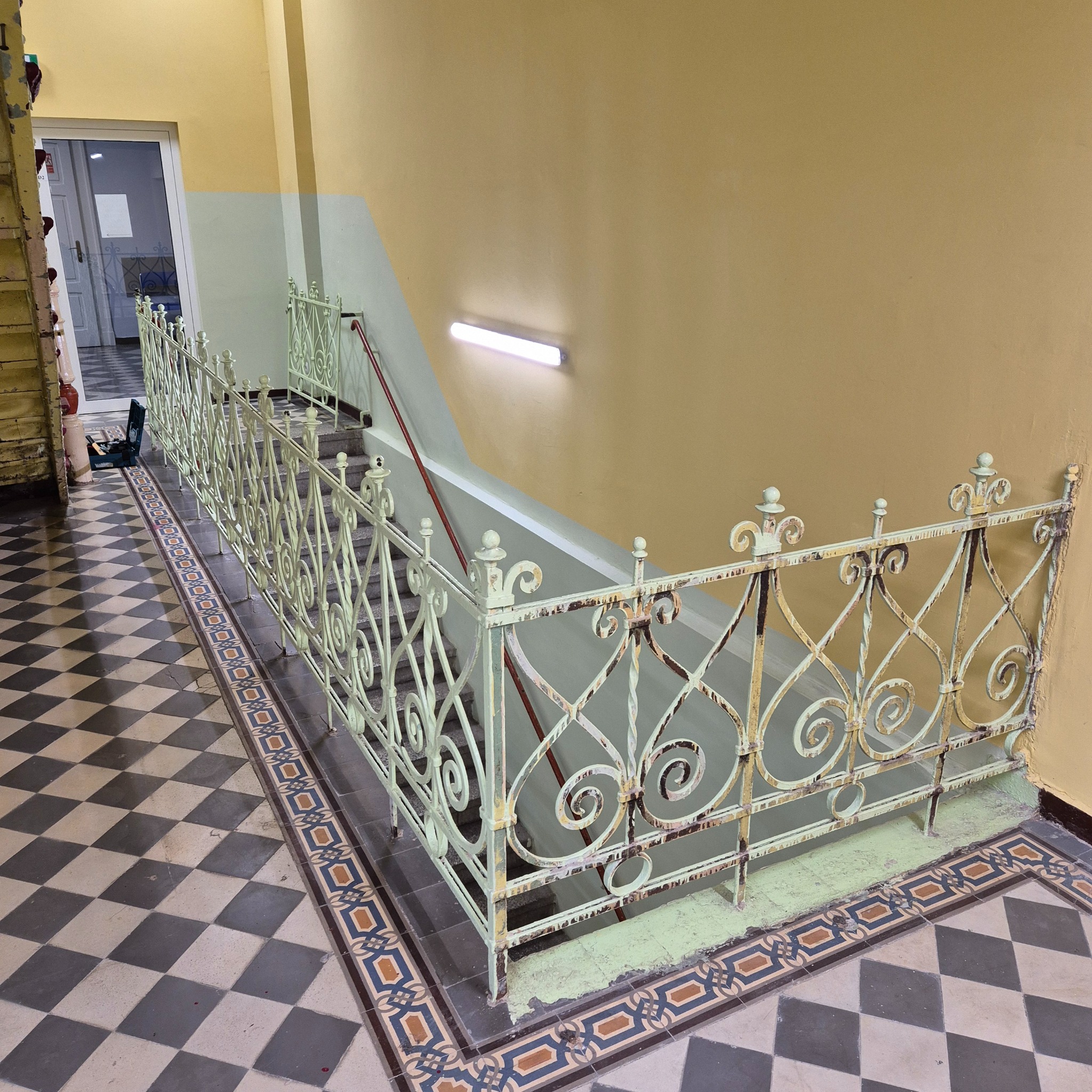 Klatka schodowa z ozdobną, metalową balustradą w stylu retro. Widoczne ślady użytkowania i odświeżona farba. Podłoga w szachownicę i narzędzia w tle.
