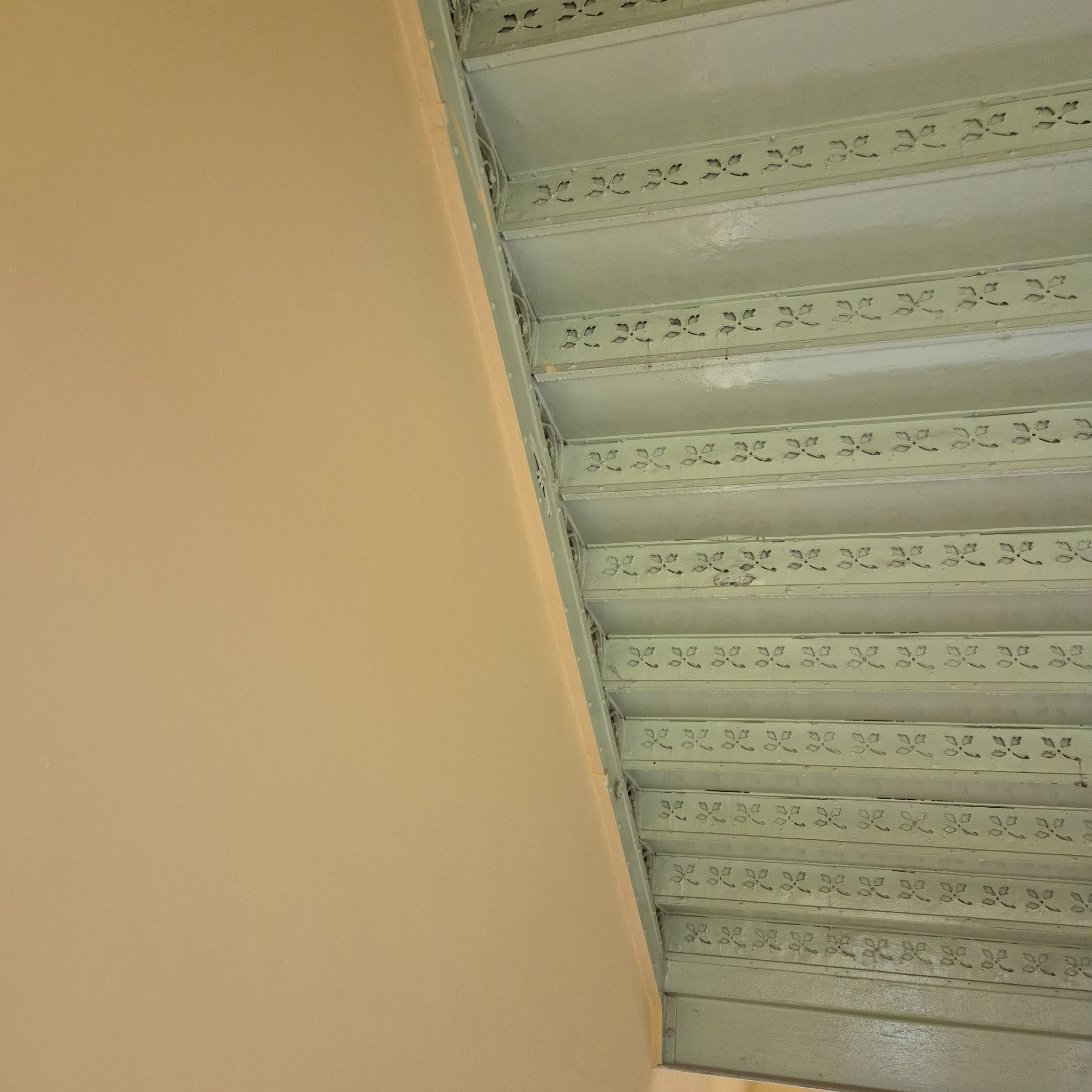 Metalowe schody z dekoracyjnym wzorem, pomalowane na jasnozielony kolor, przylegające do beżowej ściany. Widok z dołu, podkreślający detale konstrukcji.