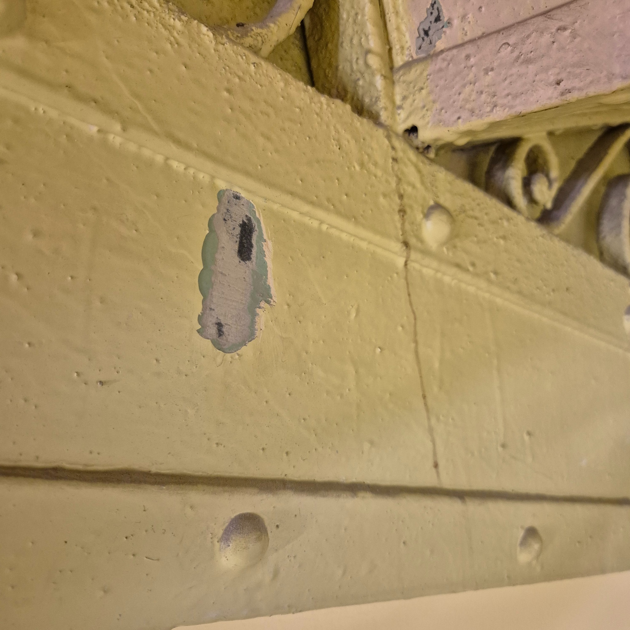 Żółta, odrapana powierzchnia z dekoracyjnym elementem. Widoczne ślady po naprawach i niedoskonałości farby. Detal architektoniczny w trakcie renowacji lub wymagający odświeżenia.