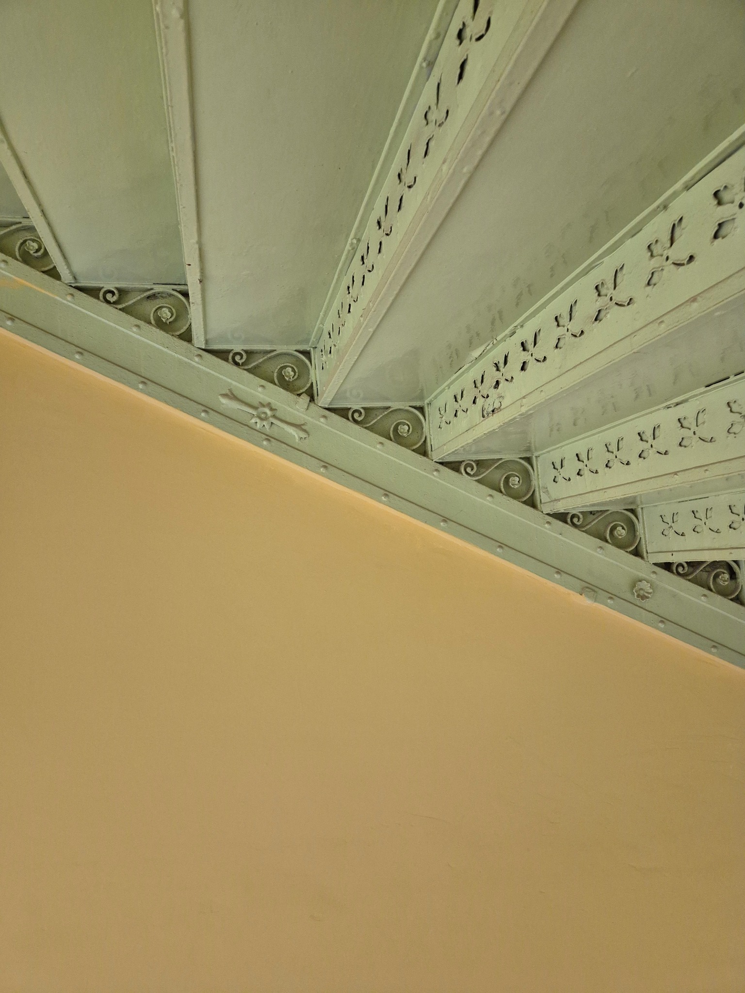 Metalowe schody z dekoracyjnymi wycięciami, widok z dołu, z fragmentem beżowej ściany. Ujęcie podkreśla detale i wzornictwo schodów.