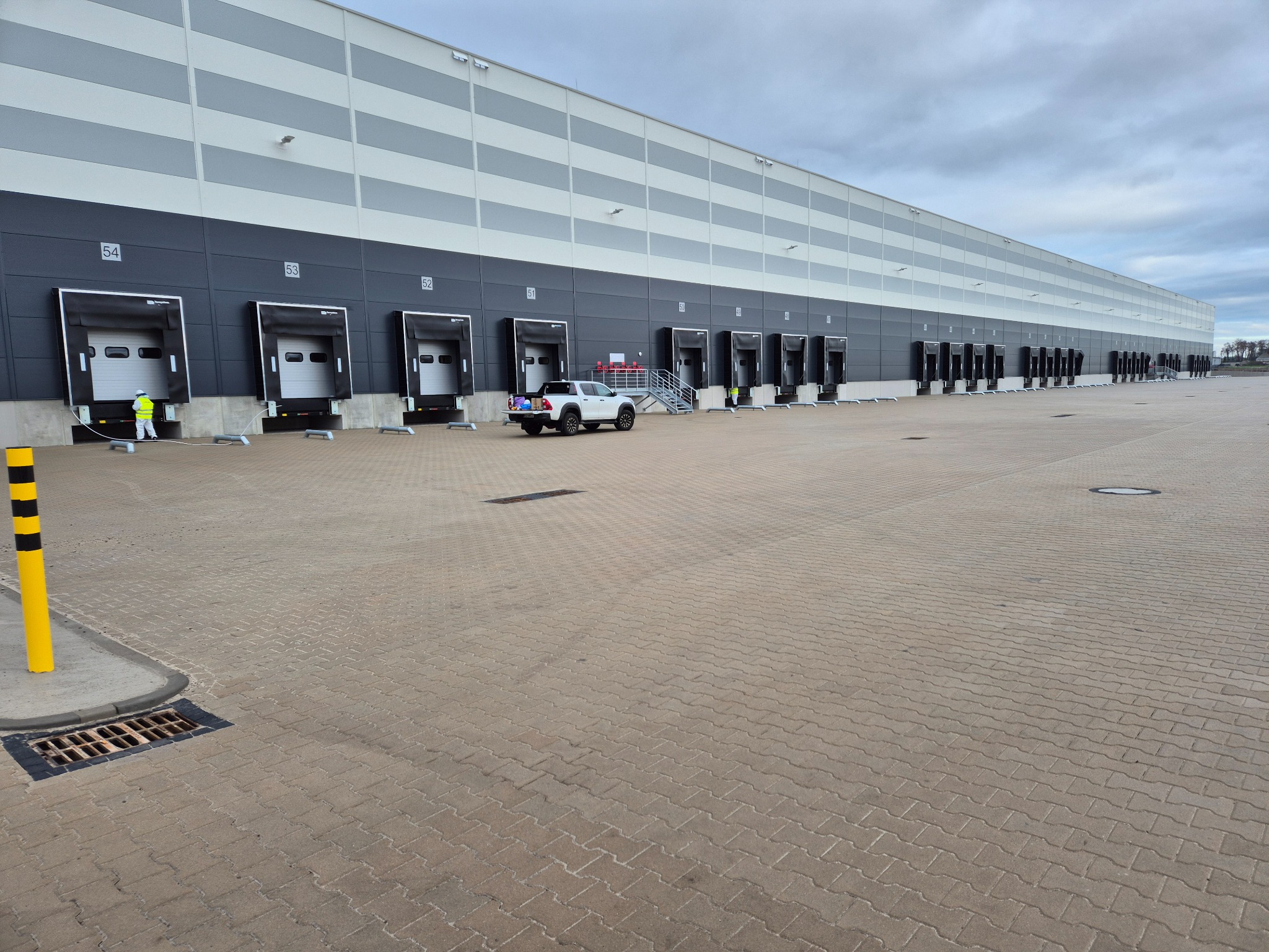 Długi budynek magazynowy z rampami załadunkowymi, biały pickup na placu. Pracownik w kamizelce przy rampie nr 54. Widok na szary plac z kostki brukowej.