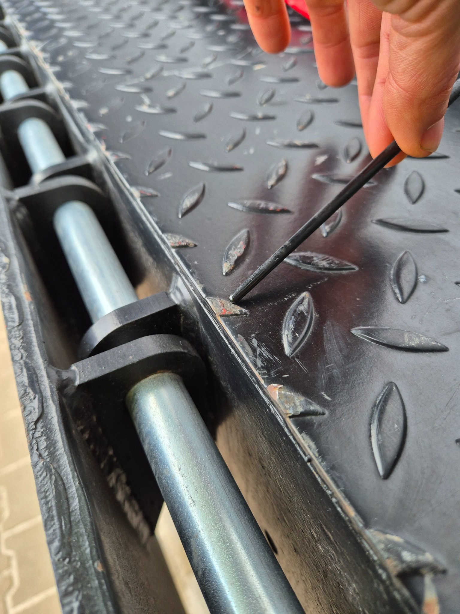 Czyszczenie metalowej konstrukcji z wzorem łezkowym. Palec wskazujący na rdzę, narzędzie w dłoni. Widoczne ślady korozji na czarnej powierzchni.