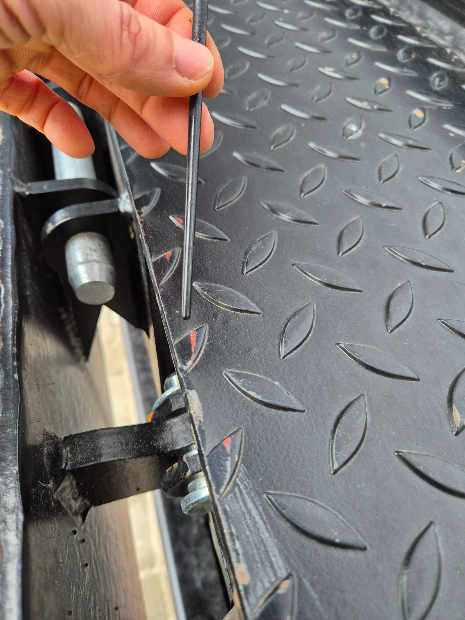 Czarny, metalowy podest z antypoślizgową fakturą, sprawdzany cienkim prętem przez dłoń. Widoczne elementy konstrukcyjne i mocowania.