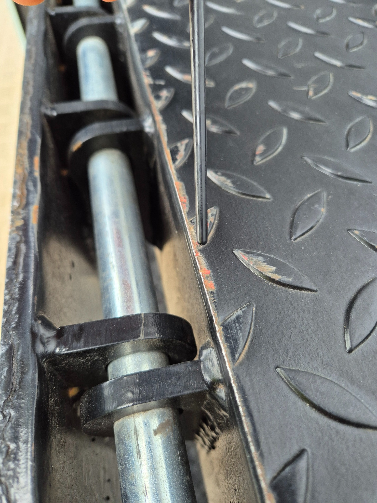 Metalowa konstrukcja z elementami antypoślizgowymi i śladami korozji, wzmocniona cylindrycznym elementem. Widoczny fragment narzędzia przy krawędzi.