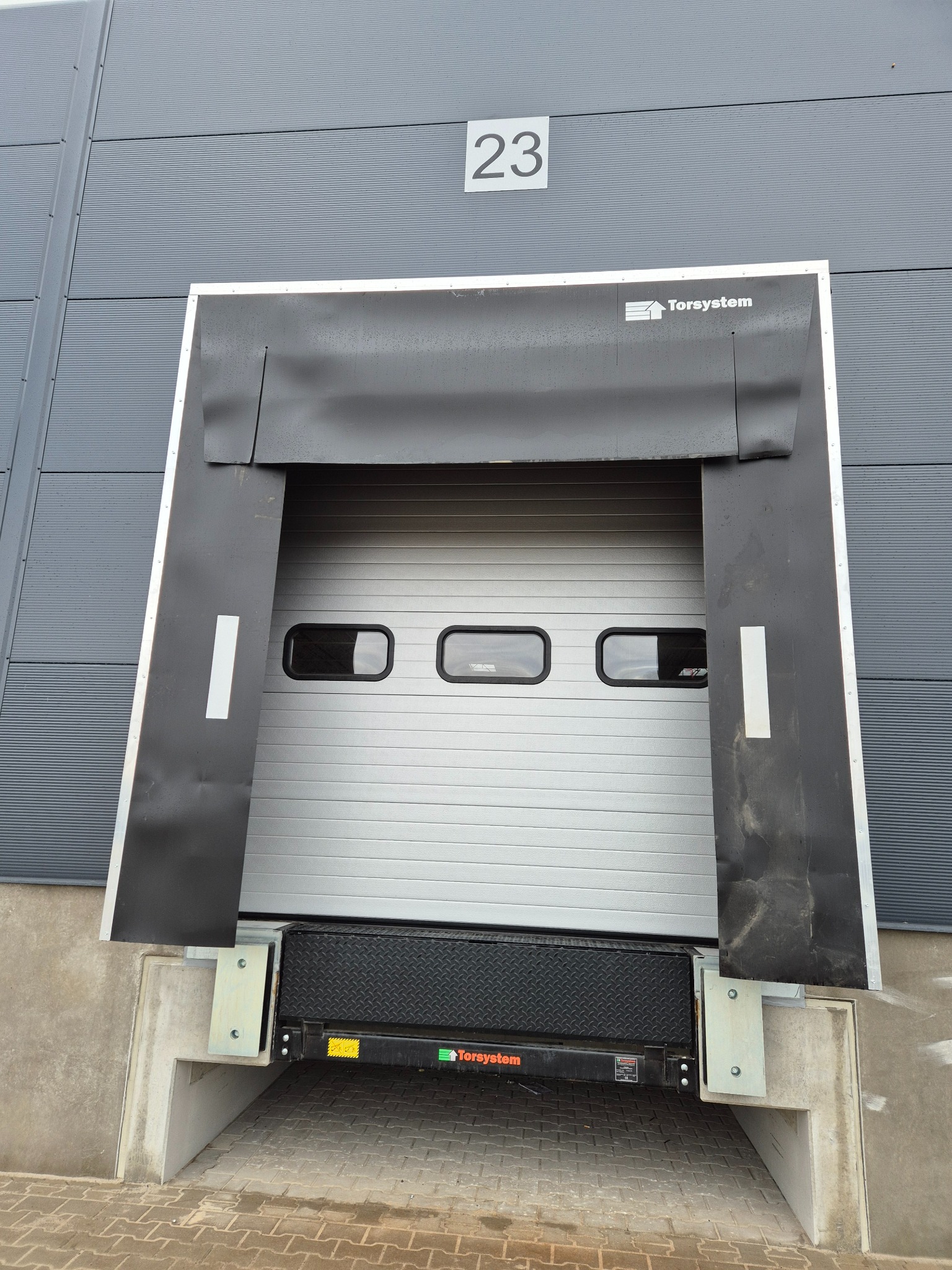 Szara brama przemysłowa Torsystem z trzema oknami, uszczelnieniem dokowym i numerem 23 na szarym budynku z blachy trapezowej. Widoczny fragment rampy załadunkowej.