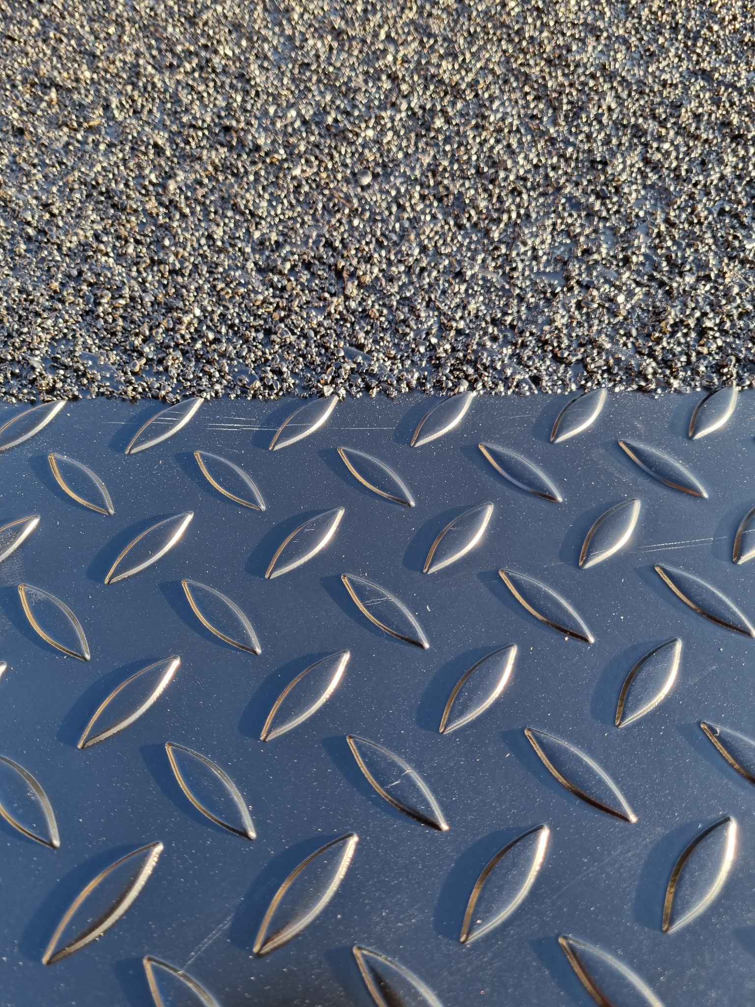 Metalowa płyta antypoślizgowa z wypukłym wzorem, częściowo pokryta szorstką, ciemną powierzchnią. Detal z bliska, ukazujący fakturę i odbicia światła.