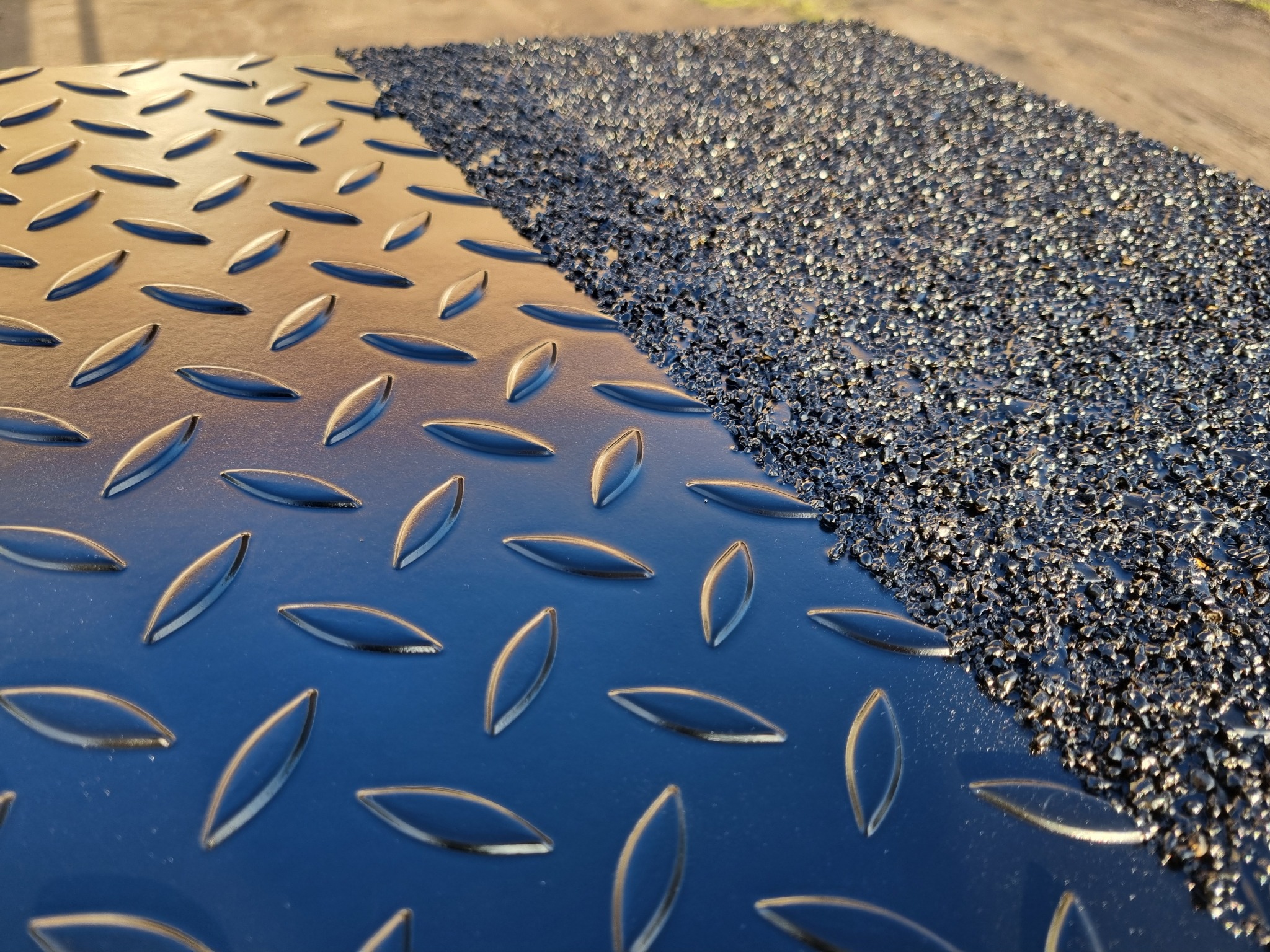 Metalowa powierzchnia z antypoślizgową powłoką, wzór łezkowy. Część powierzchni pokryta drobnym, czarnym granulatem. Kontrast faktur i połysku.