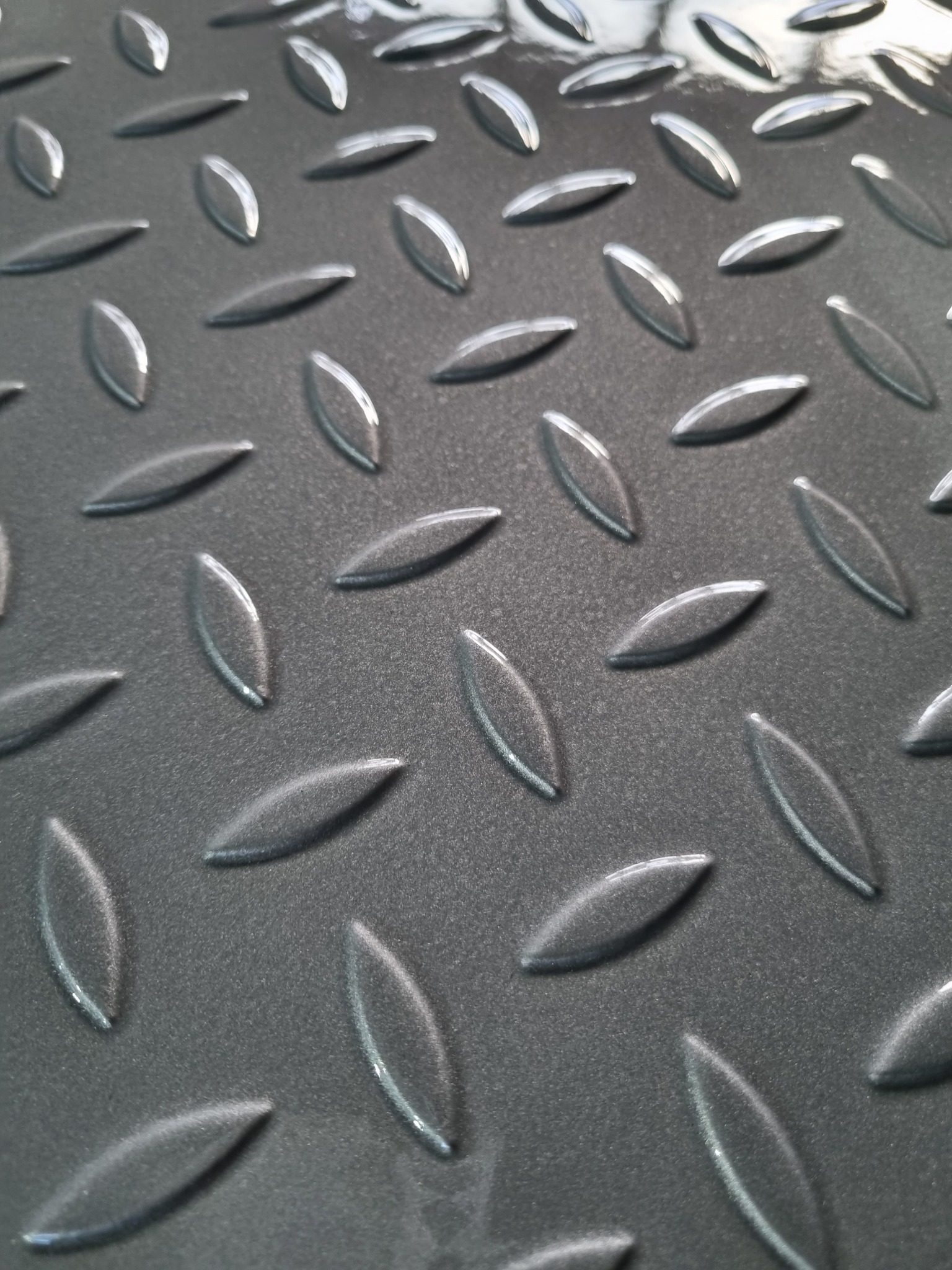 Szara, metalowa powierzchnia z wytłoczonym wzorem w kształcie łezek, ułożonych ukośnie. Błyszcząca faktura, widoczne refleksy światła. Detal metalowej podłogi.