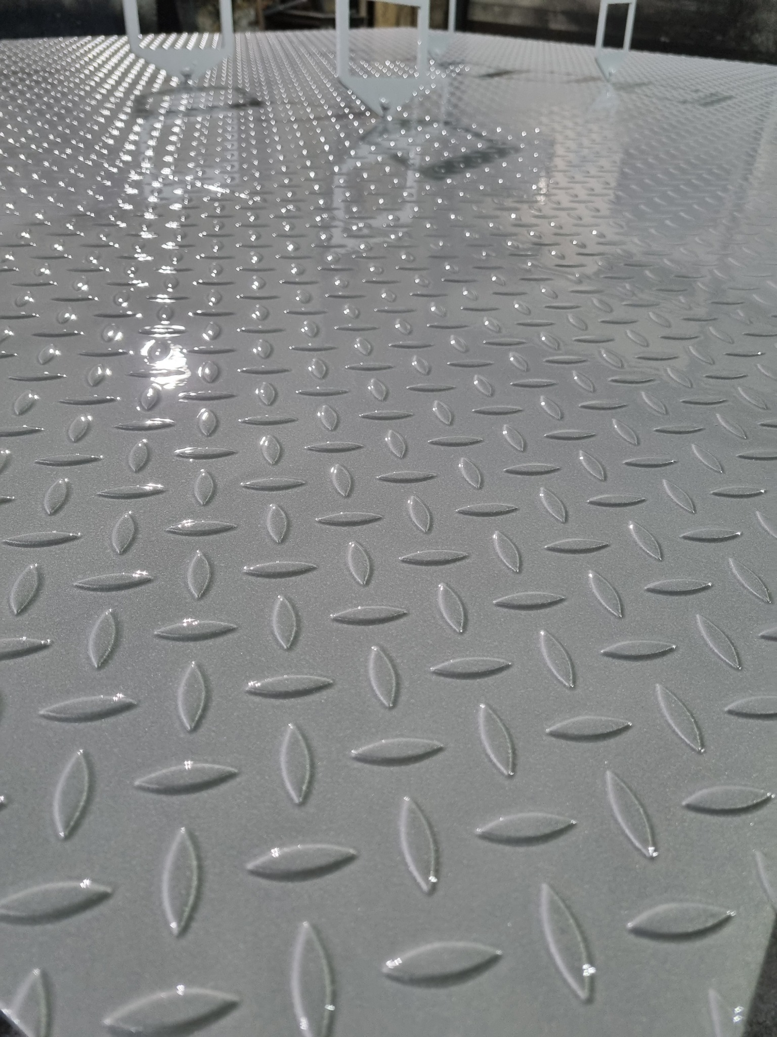 Błyszcząca, szara, metalowa powierzchnia z wytłoczonym wzorem łezkowym, prawdopodobnie po świeżym malowaniu. Widoczne odbicia światła.