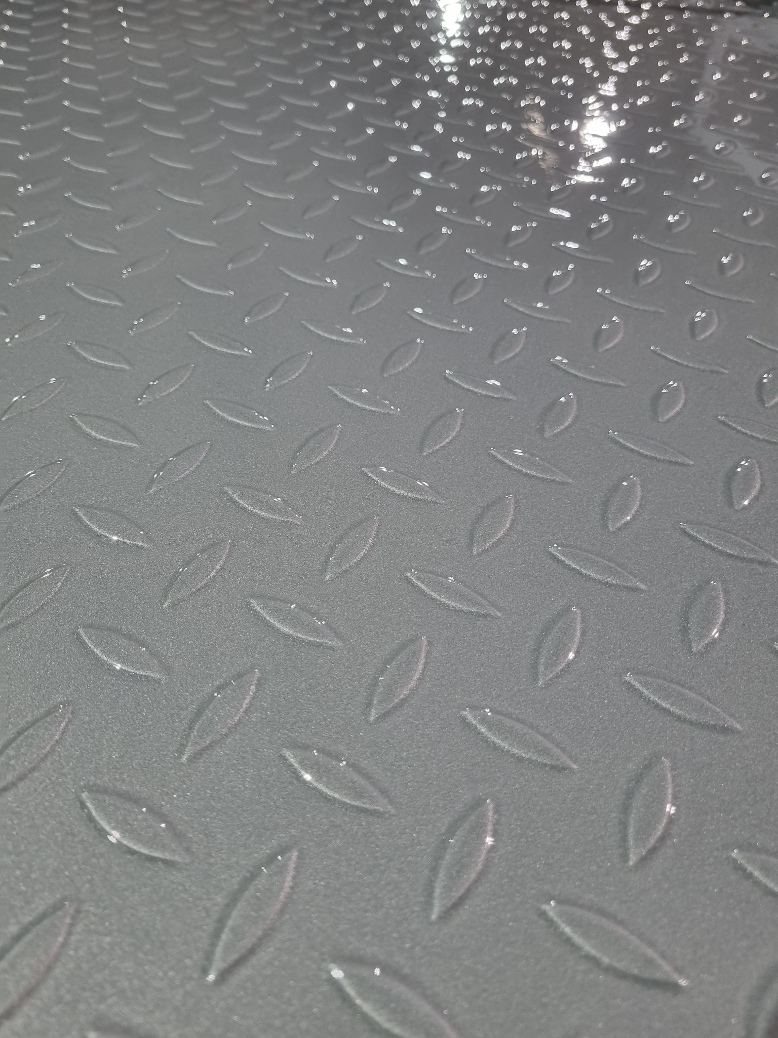 Szara, metalowa powierzchnia z wytłoczonym wzorem łezkowym, lekko odbijająca światło. Widoczne drobne niedoskonałości powłoki, sugerujące proces piaskowania lub konserwacji.