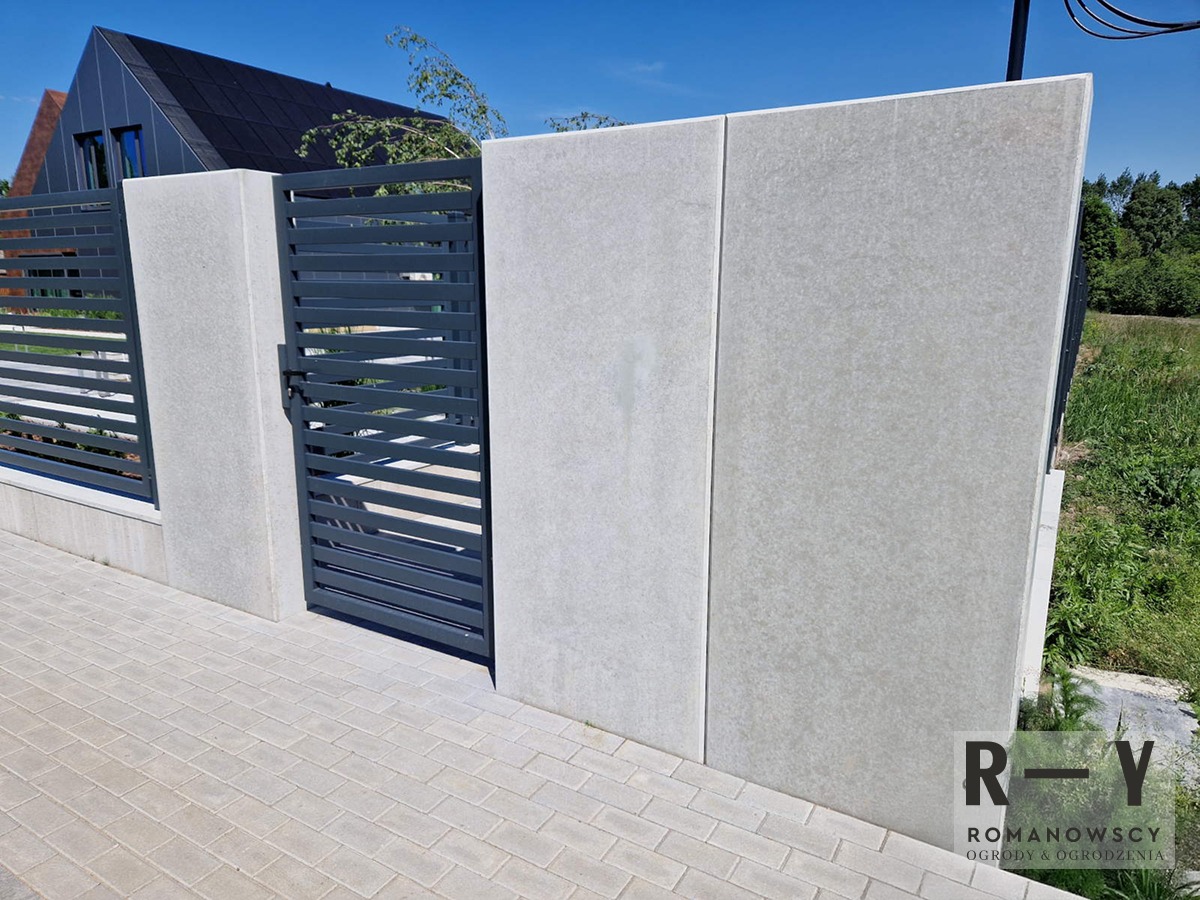 Nowoczesne ogrodzenie z betonowych paneli i metalowych przęseł, z bramką, na tle kostki brukowej i domu z ciemną dachówką, w słoneczny dzień.