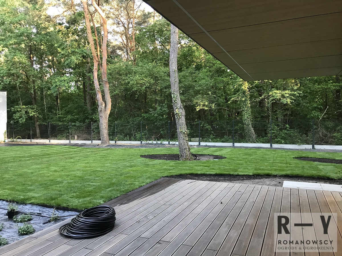 Nowo założony trawnik z rolki, z widocznymi obrzeżami i systemem nawadniania, otoczony zielenią drzew, z drewnianym tarasem w pierwszym planie.