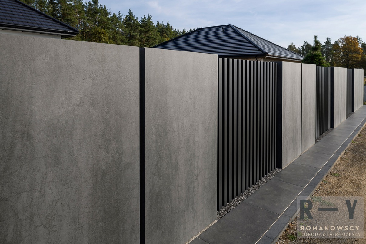 Nowoczesne ogrodzenie panelowe z betonu architektonicznego i czarnych, pionowych listew, oddzielające posesję od otoczenia. Minimalistyczny design w szarościach.