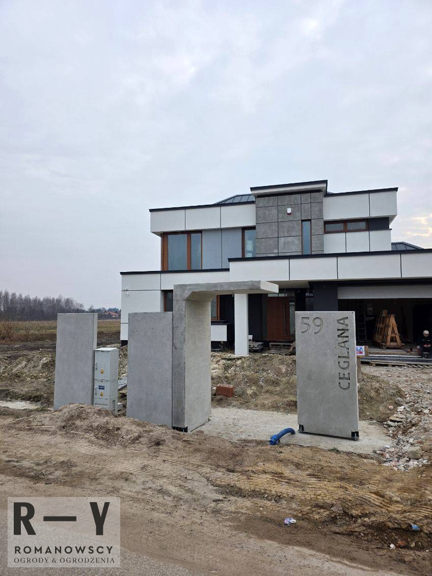 Nowoczesna brama wejściowa z betonu architektonicznego przed domem, z numerem '59 Ceglana'. W tle widoczny budynek w trakcie prac wykończeniowych.