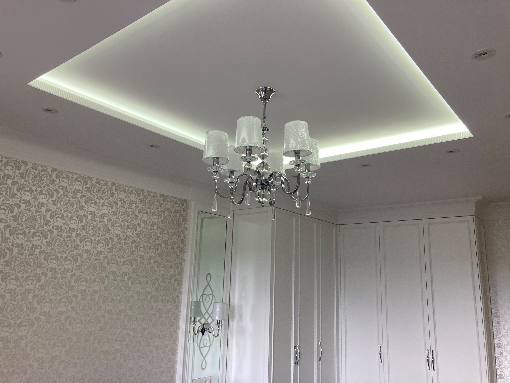 Sufit podwieszany z oświetleniem LED i ozdobnym żyrandolem w eleganckim wnętrzu z tapetą w kwiaty i białą zabudową meblową.
