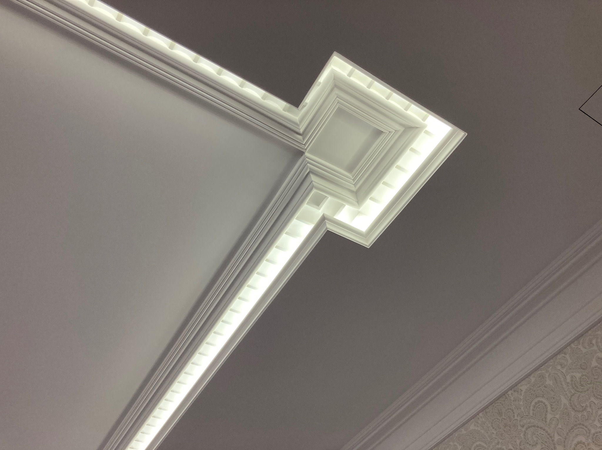 Dekoracyjny sufit z oświetleniem LED w narożniku, widok pod kątem. Białe listwy sufitowe tworzą geometryczny wzór z subtelnym podświetleniem.