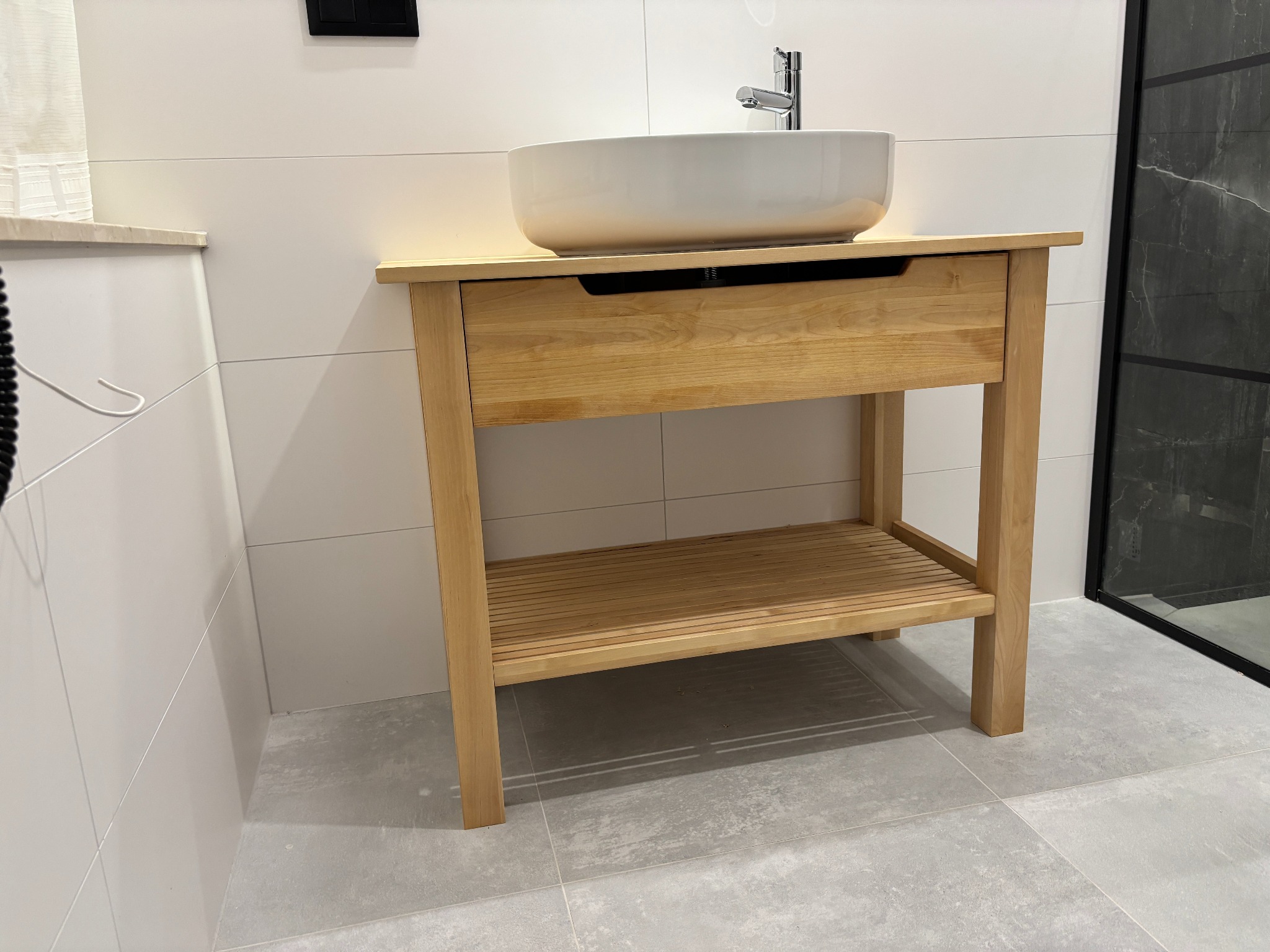 Jasna, drewniana szafka pod umywalkę z litego drewna brzozowego, olejowana, z szufladą i półką, na tle białych płytek i szarej podłogi.
