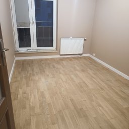Phu Erwin Franek - Jasna, odnowiona podłoga parkietowa w pustym pokoju z oknem i grzejnikiem. Widoczne listwy przypodłogowe i fragment drzwi. Parkiet ułożony w cegiełkę.