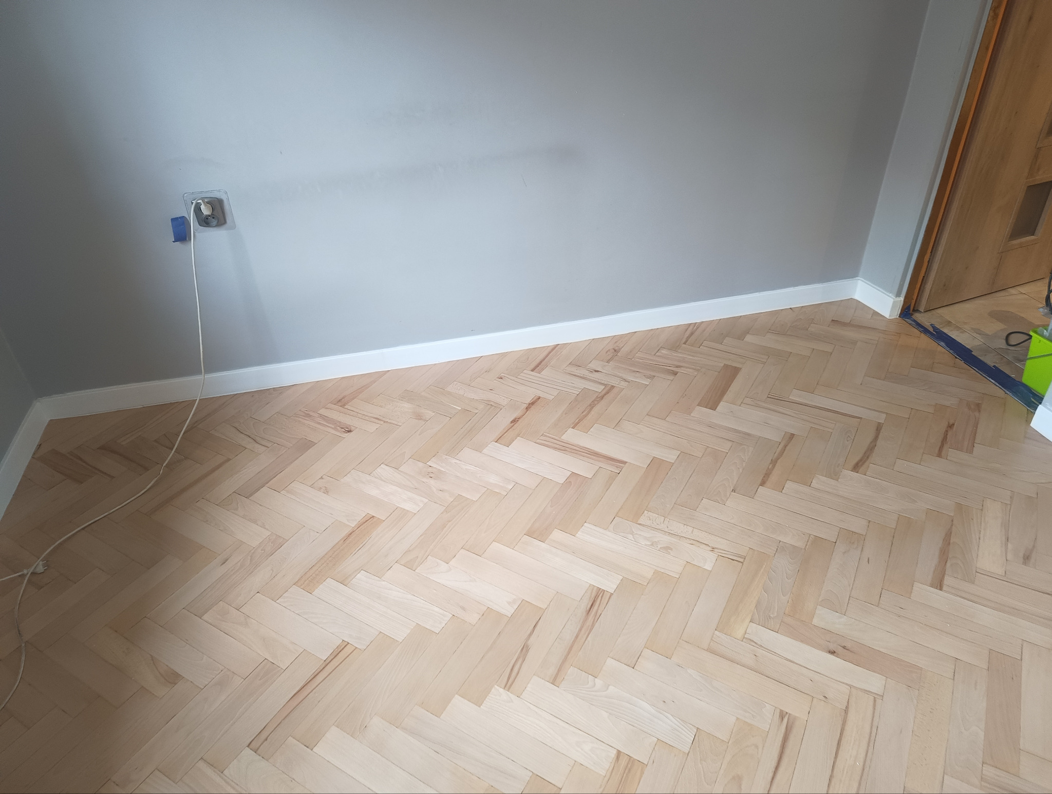 Jasny parkiet ułożony w jodełkę w pokoju z szarą ścianą i białą listwą przypodłogową. Widoczne gniazdko elektryczne i fragment drzwi.