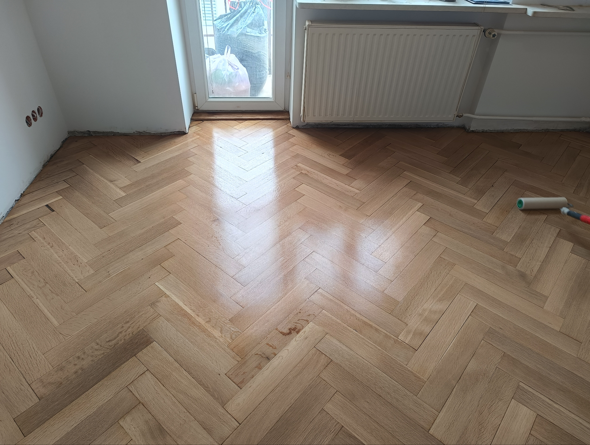 Parkiet ułożony w jodełkę, świeżo po cyklinowaniu i lakierowaniu. Widoczny wałek malarski, okno i grzejnik. Jasne drewno, połyskująca powierzchnia.