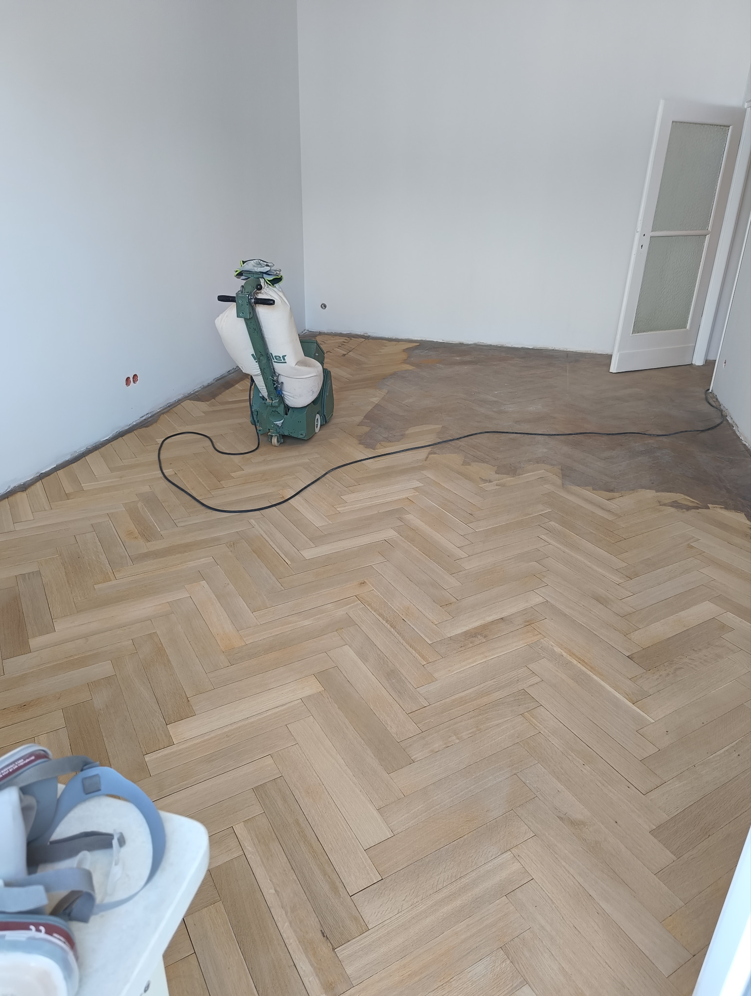 Cyklinowanie parkietu w jodełkę: maszyna do cyklinowania w trakcie pracy, widoczna różnica w kolorze między obszarem obrobionym a surowym drewnem. Białe ściany i drzwi w tle.