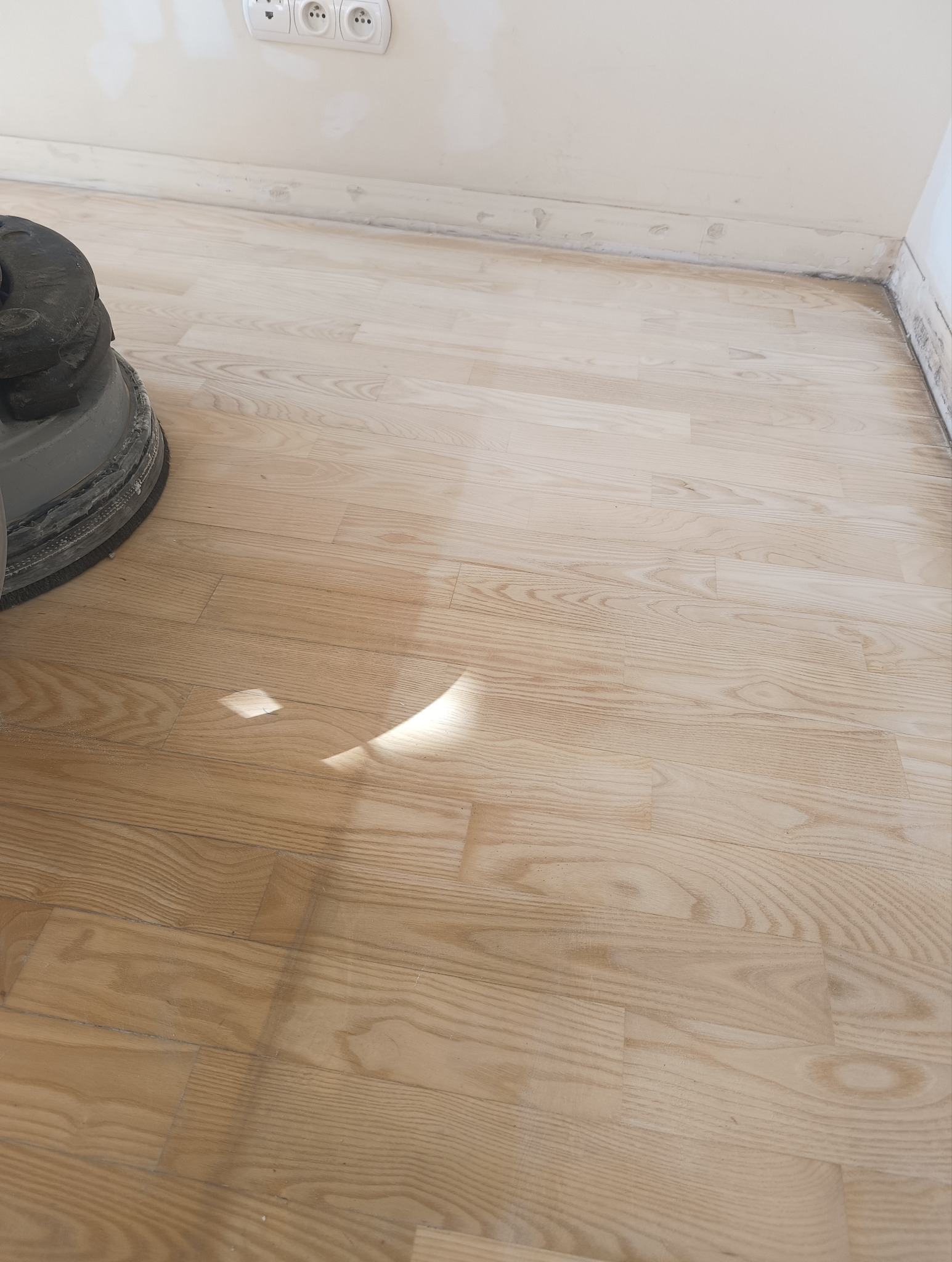 Jasna, odnowiona podłoga parkietowa z widoczną maszyną do cyklinowania w rogu. Świeżo wyszlifowana powierzchnia, gotowa do dalszej obróbki.