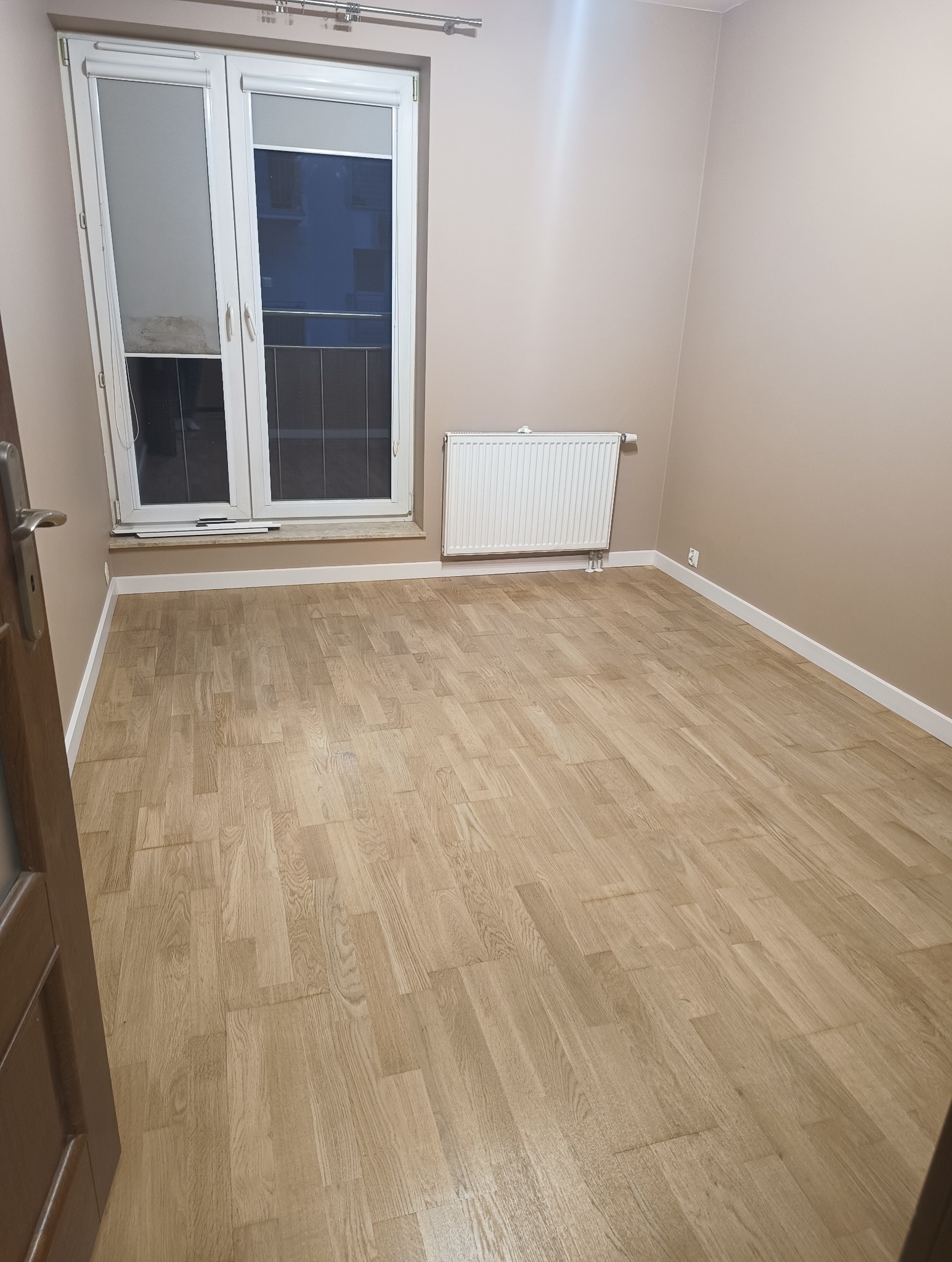 Jasna, odnowiona podłoga parkietowa w pustym pokoju z oknem i grzejnikiem. Widoczne listwy przypodłogowe i fragment drzwi. Parkiet ułożony w cegiełkę.