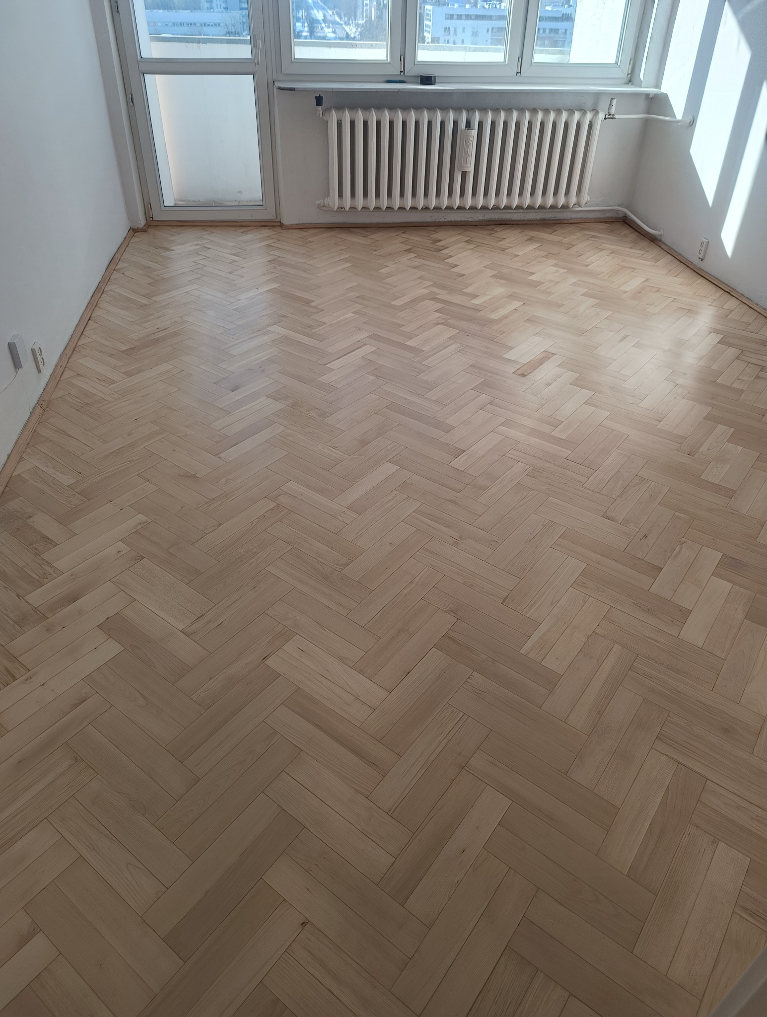 Jasna, odnowiona podłoga parkietowa ułożona w jodełkę francuską, widok z góry, z grzejnikiem i oknem w tle. Świeżo położony lakier podkreśla naturalny rysunek drewna.