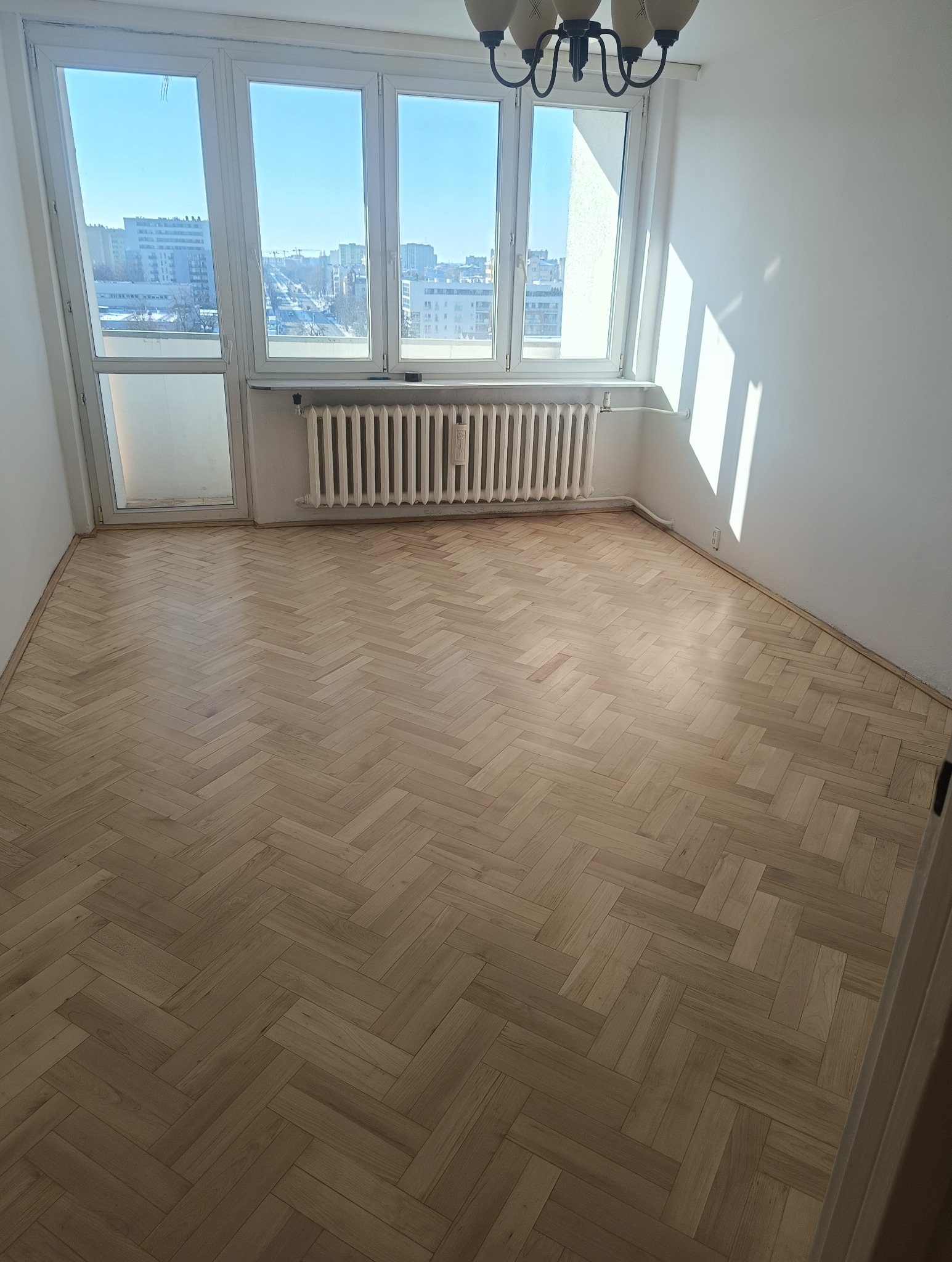 Jasna, odnowiona podłoga parkietowa w jodełkę, po cyklinowaniu. Widok na okno z widokiem na miasto. Grzejnik pod oknem. Świeże, jasne wnętrze po renowacji.