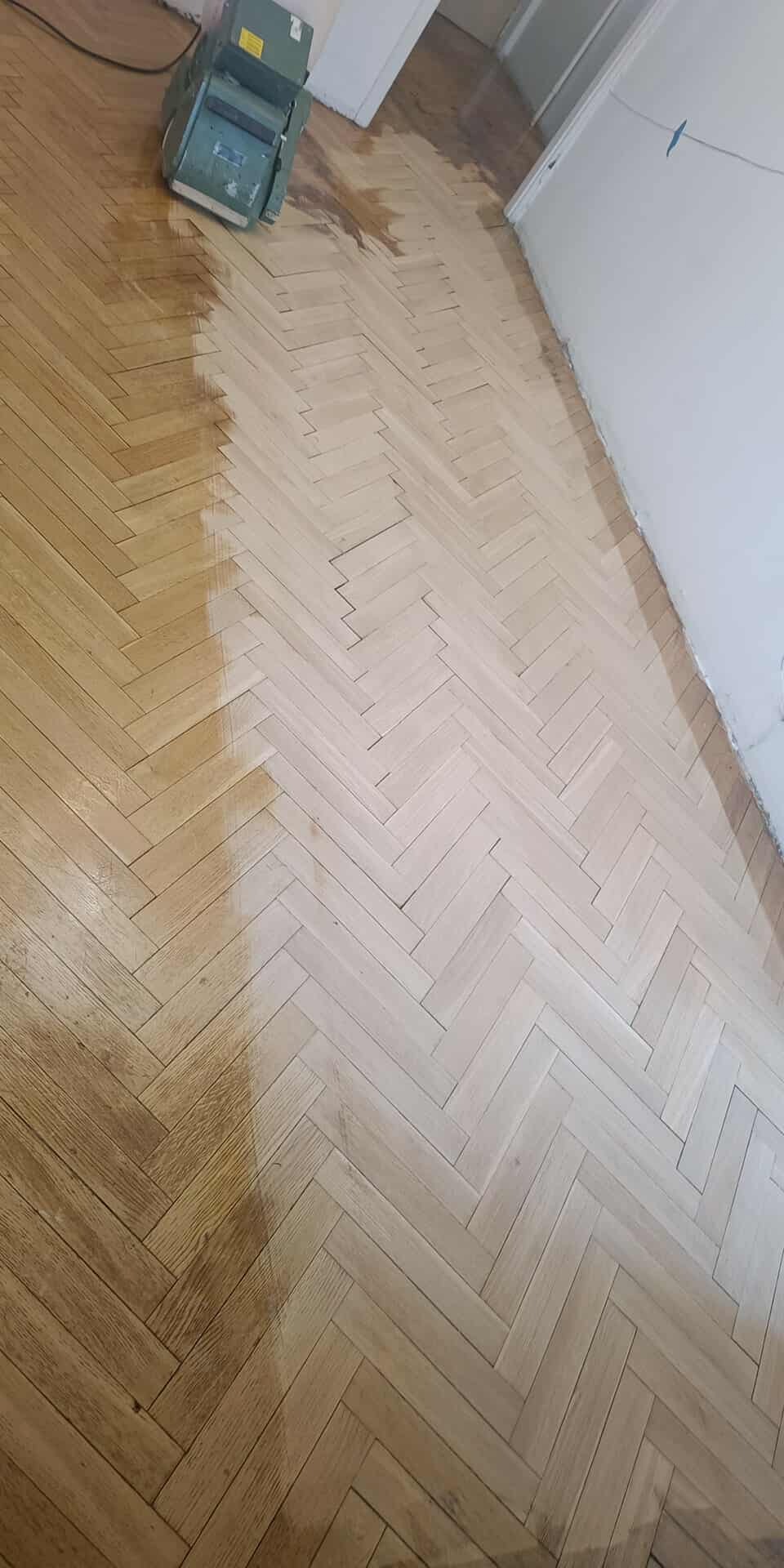 Parkiet w jodełkę częściowo odnowiony, z widoczną różnicą w kolorze drewna po zabiegu. Szlifierka do parkietu w rogu kadru. Widok z góry.