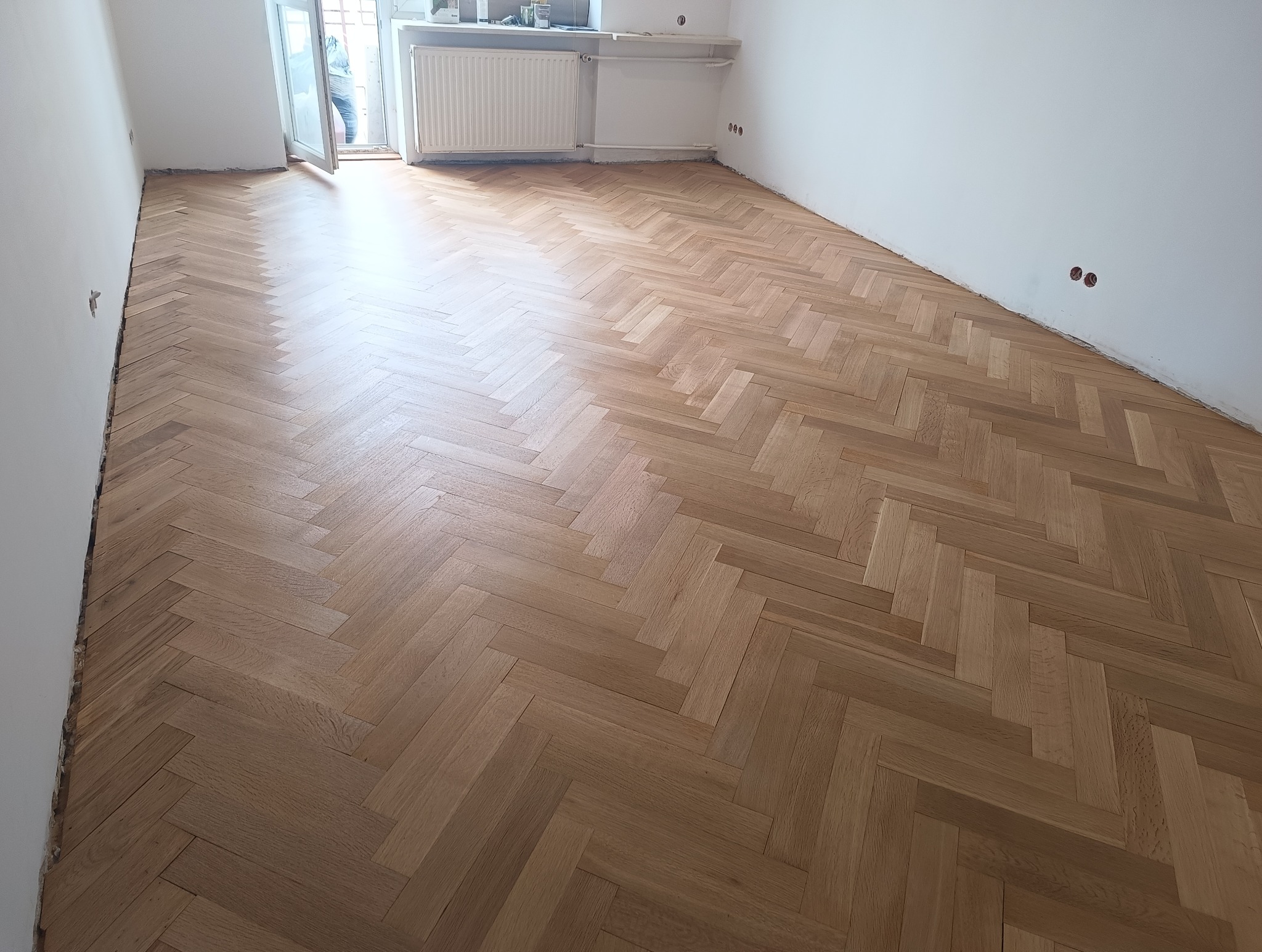 Parkiet ułożony w jodełkę po renowacji, jasne drewno, widoczne ślady po szlifowaniu przy ścianach, w tle okno i grzejnik. Świeżo wycyklinowana podłoga.